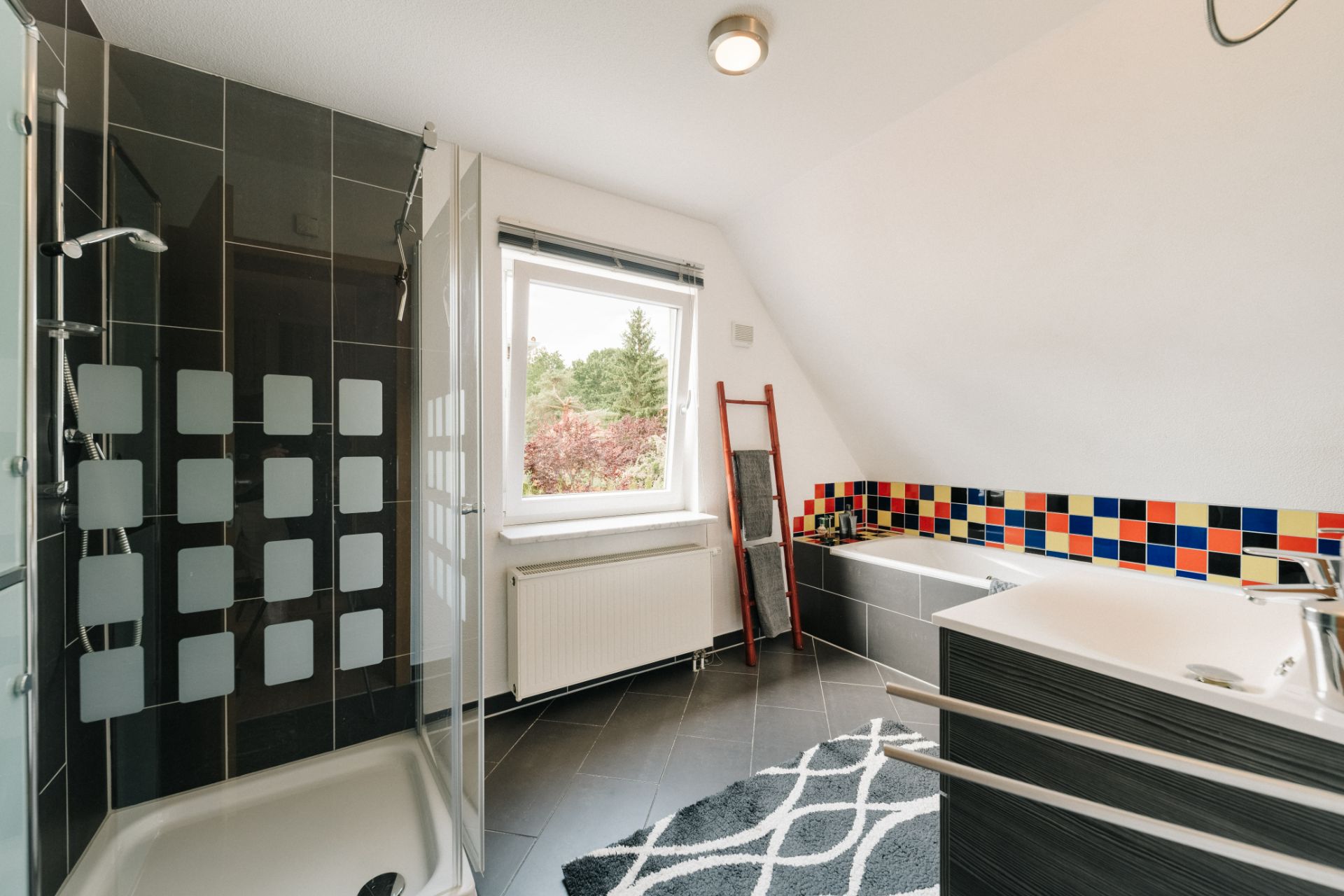 Modernes Badezimmer mit Dusche, Badewanne, Fenster und farbigen Fliesen an der Wand.