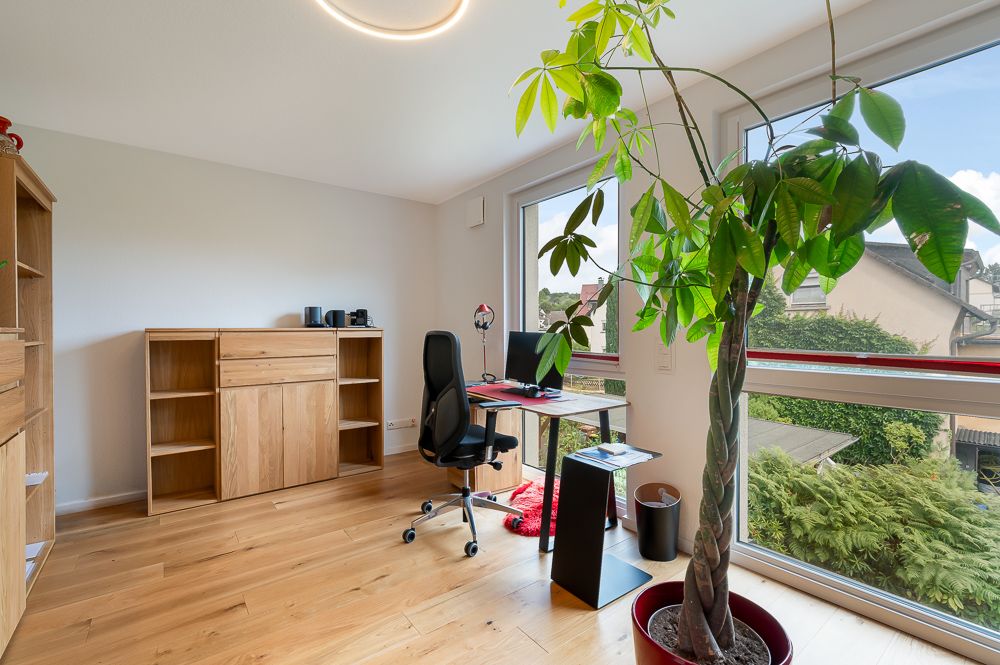 Helles Arbeitszimmer mit großem Fenster, Schreibtisch, Stuhl, Holzregal und großer Zimmerpflanze.