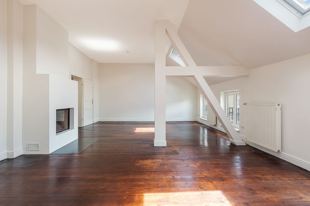 Helles, leeres Zimmer mit Holzboden, Kamin, sichtbaren Balken und großen Fenstern.