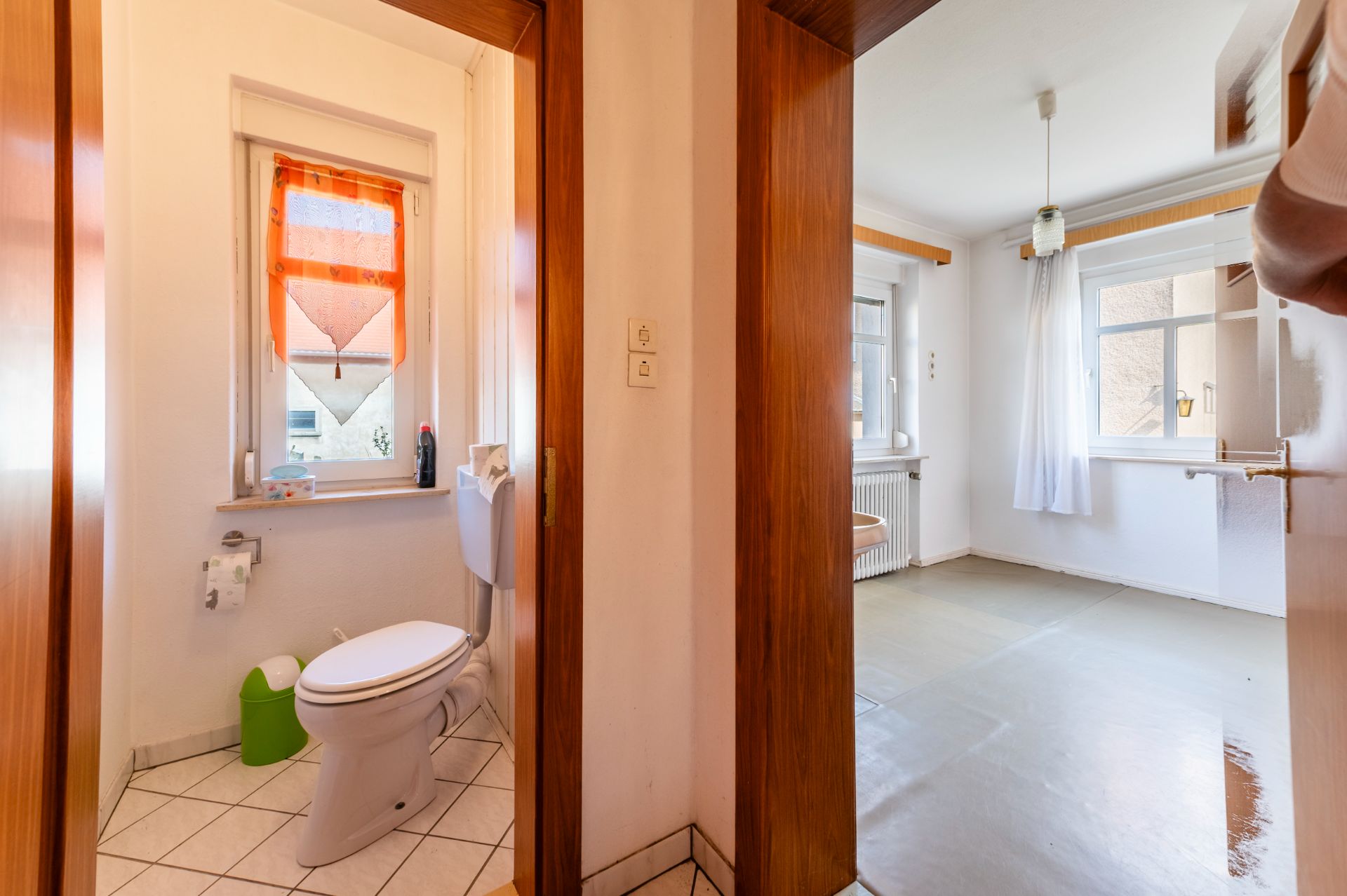 Blick vom Flur auf ein Badezimmer mit Toilette und Fenster sowie in ein helles, leeres Zimmer.