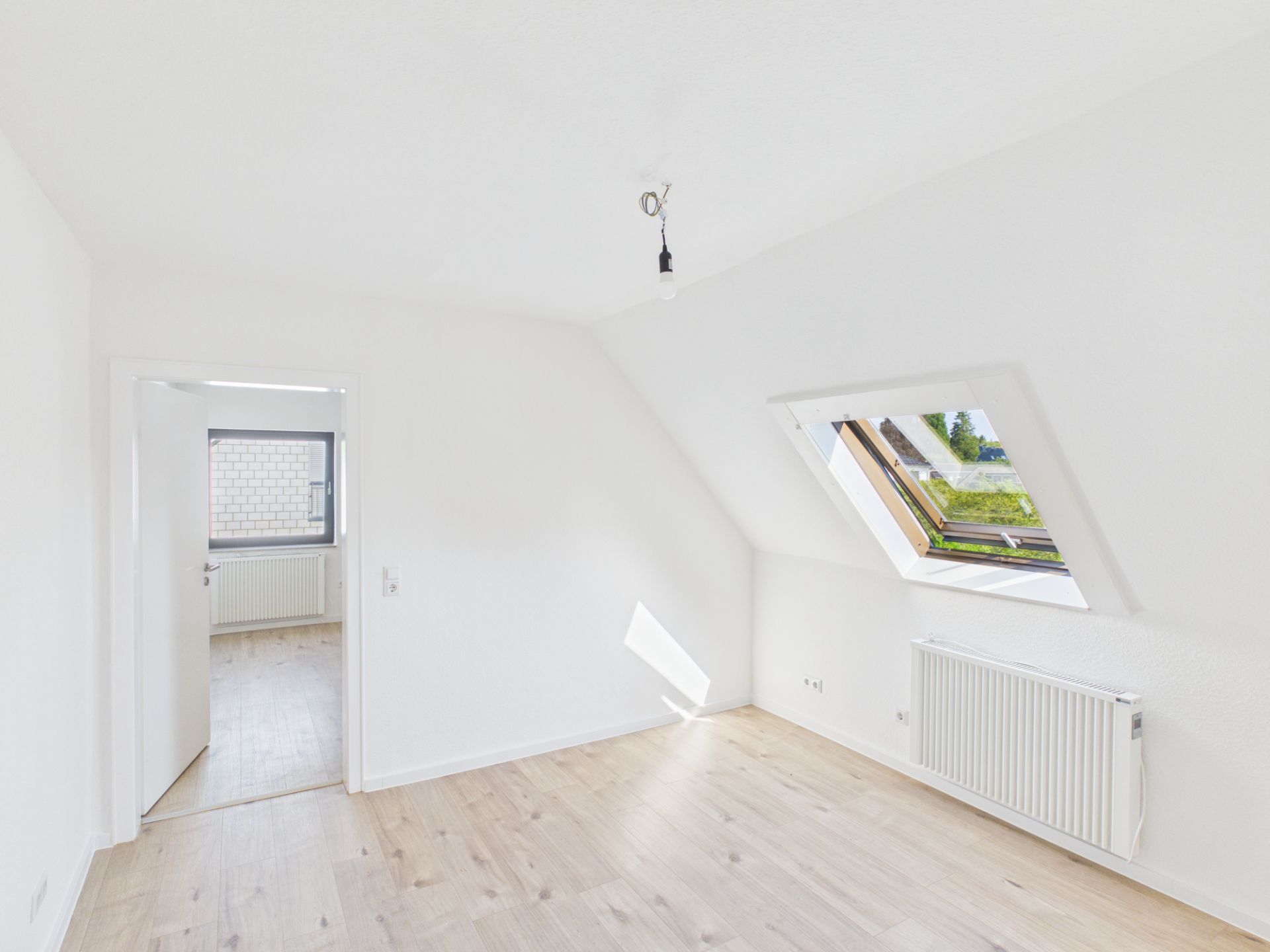 Helles Zimmer mit schräger Decke, Dachfenster und Holzboden, Tür führt in einen weiteren Raum.
