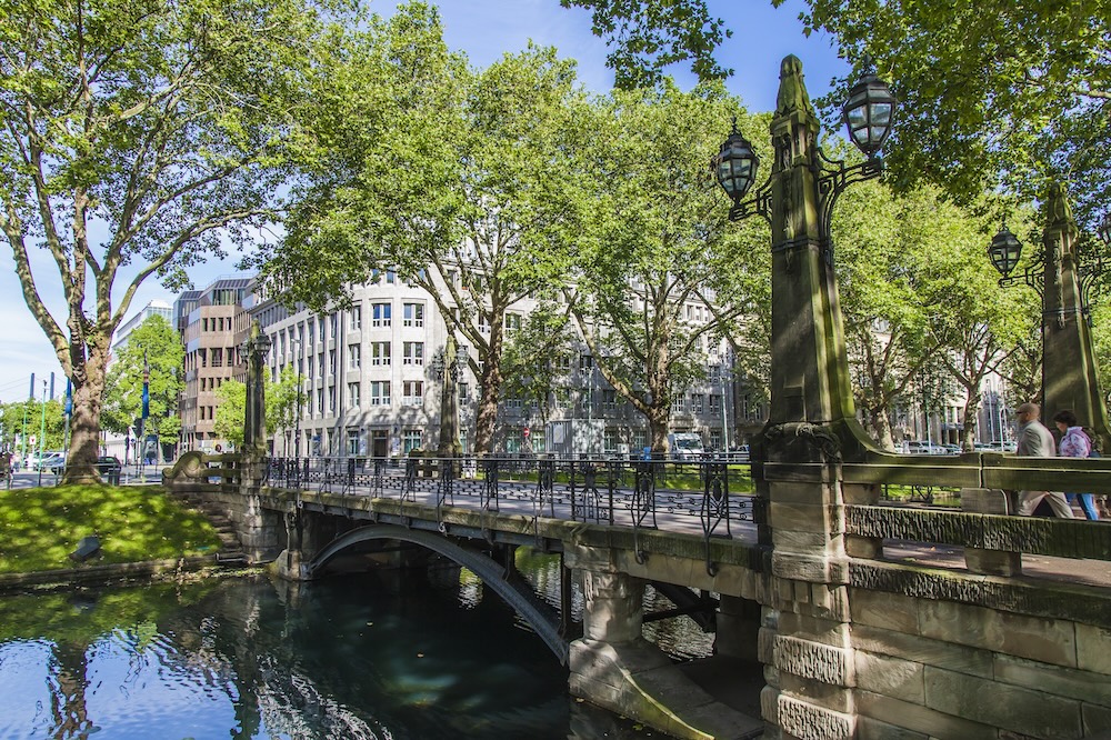 Steinerne Brücke über einen Kanal, umgeben von Bäumen und Gebäuden im Hintergrund.