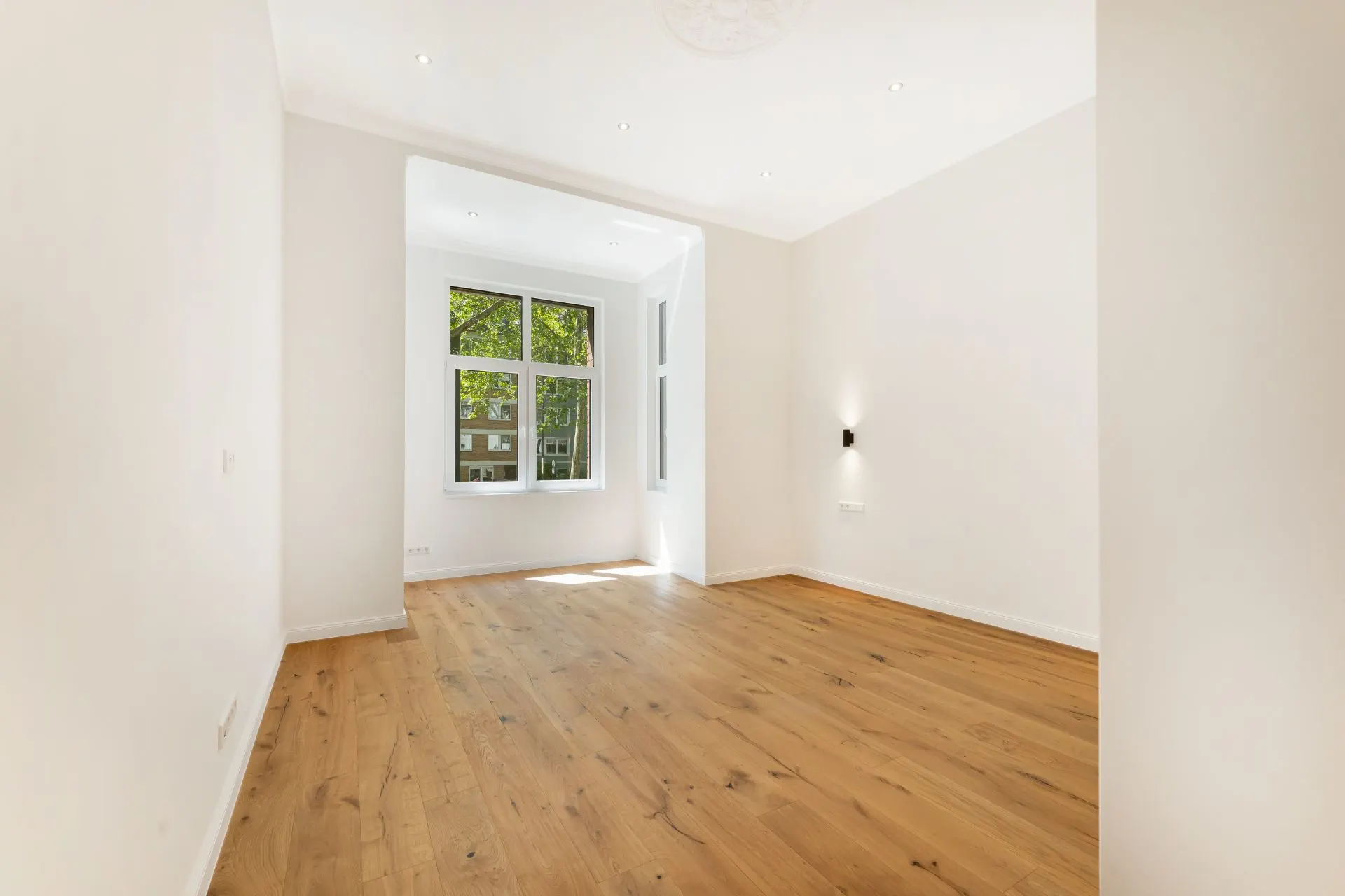 Helles, leeres Zimmer mit Holzfußboden und großem Fenster.