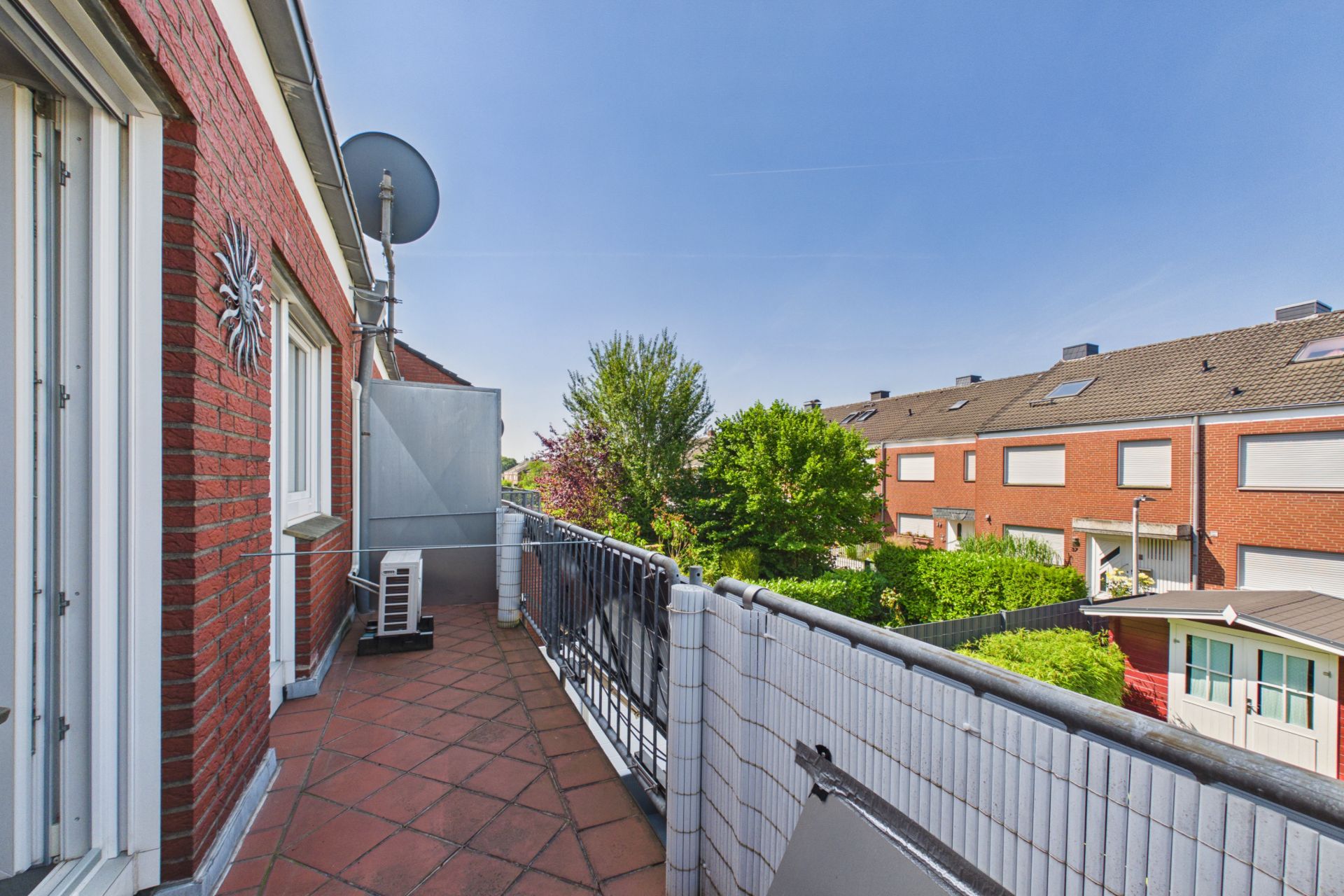 Balkon mit rotem Fliesenboden, Blick auf Nachbarhäuser und grüne Bäume.