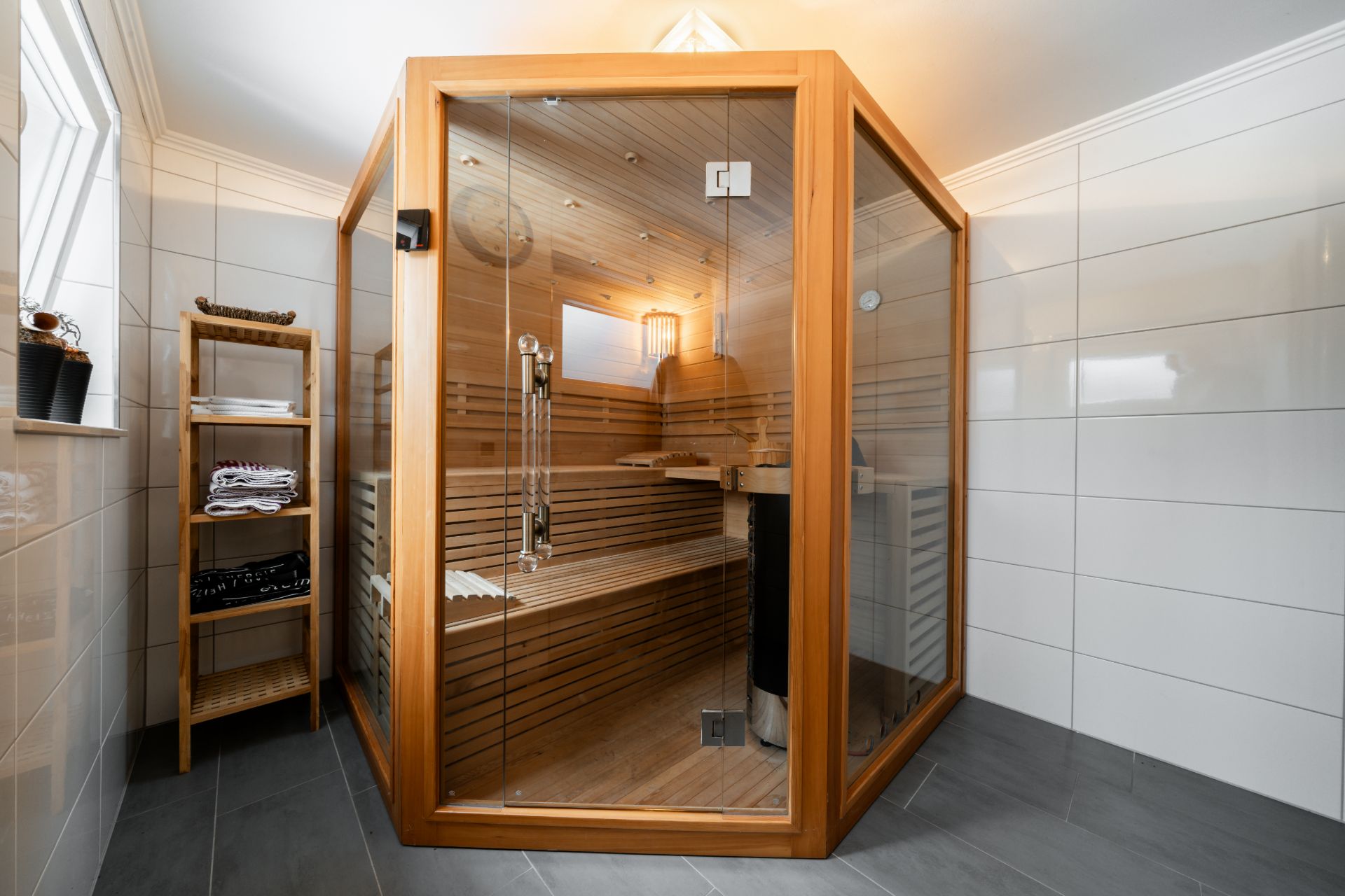 Moderne Heimsauna mit Glaswänden in einem gefliesten Raum, daneben ein Regal mit Handtüchern.