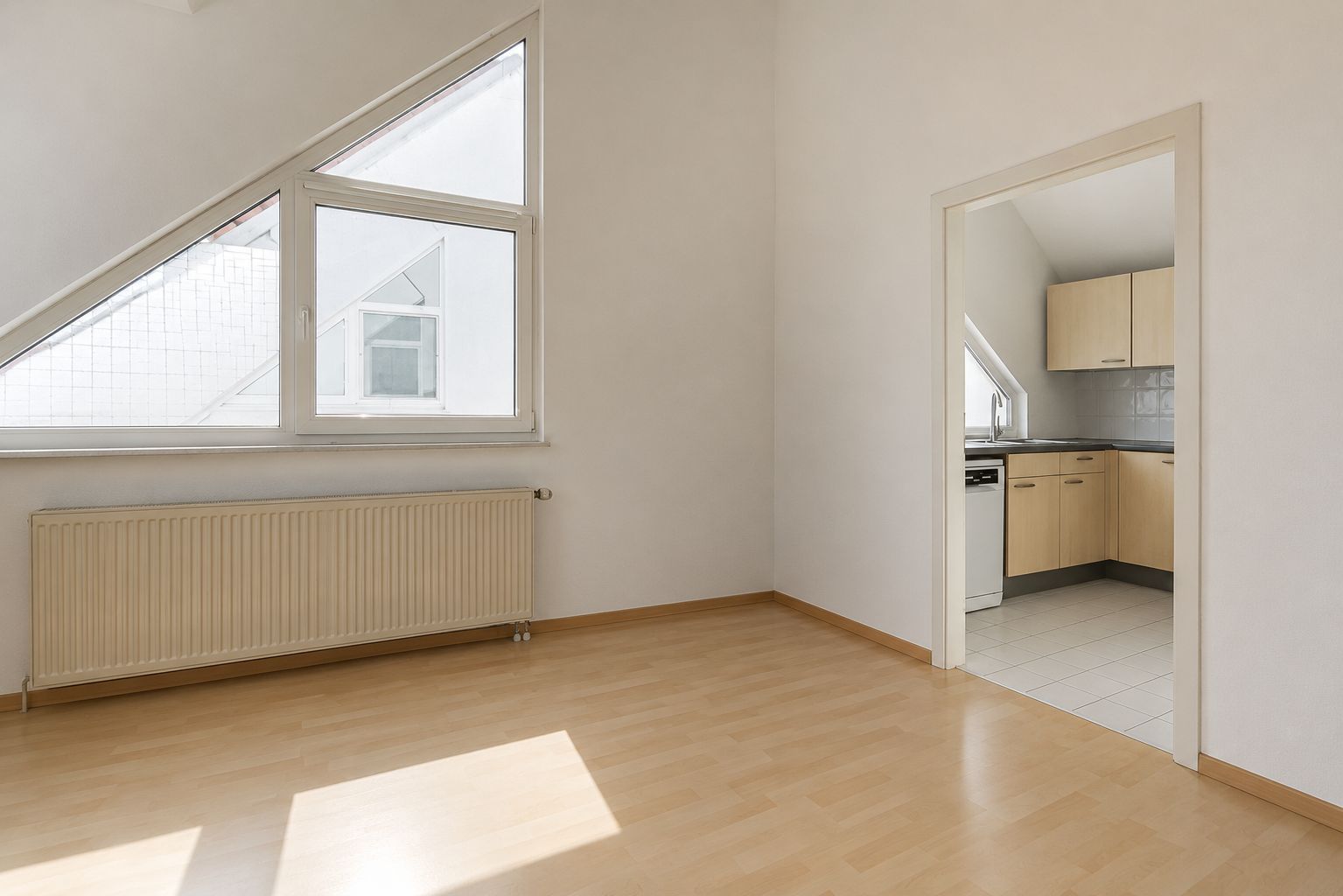 Helles Zimmer mit schrägem Fenster, Heizkörper und Durchgang zur Küche mit Einbauschränken.