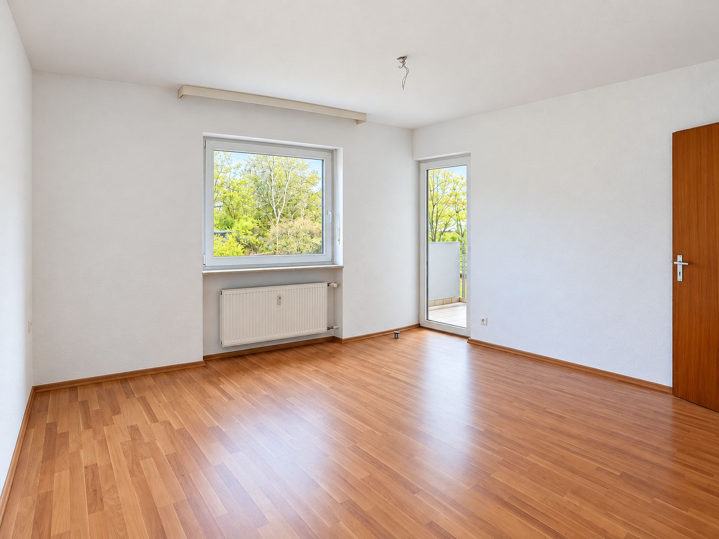 Leeres Zimmer mit Holzfußboden, großem Fenster, Tür zum Balkon und weiß gestrichenen Wänden.