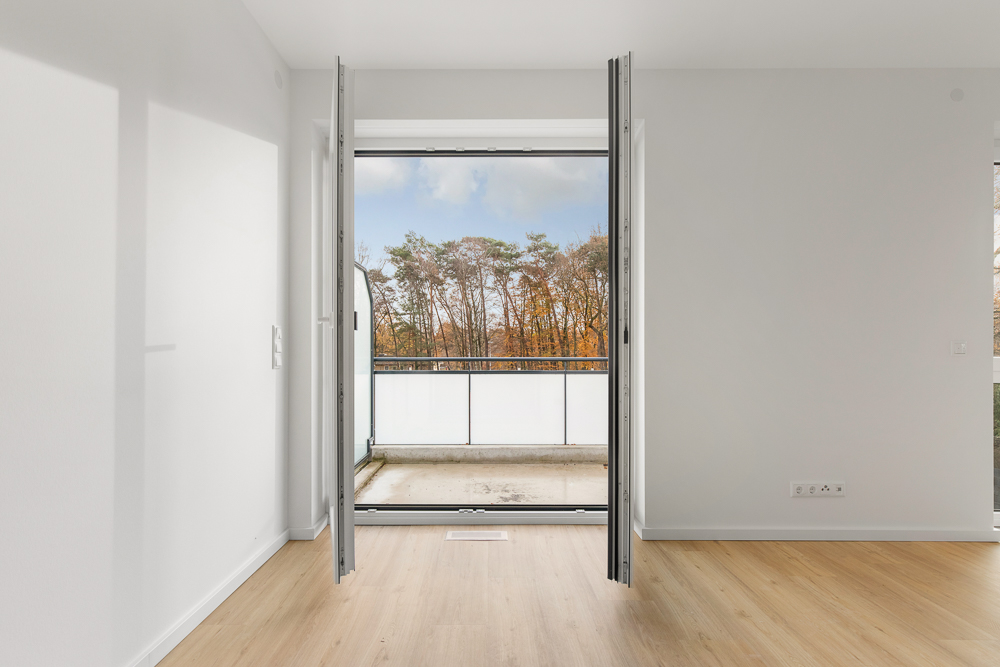 Helles Zimmer mit Holzfußboden und offener Tür zu einem Balkon mit Ausblick auf Bäume.