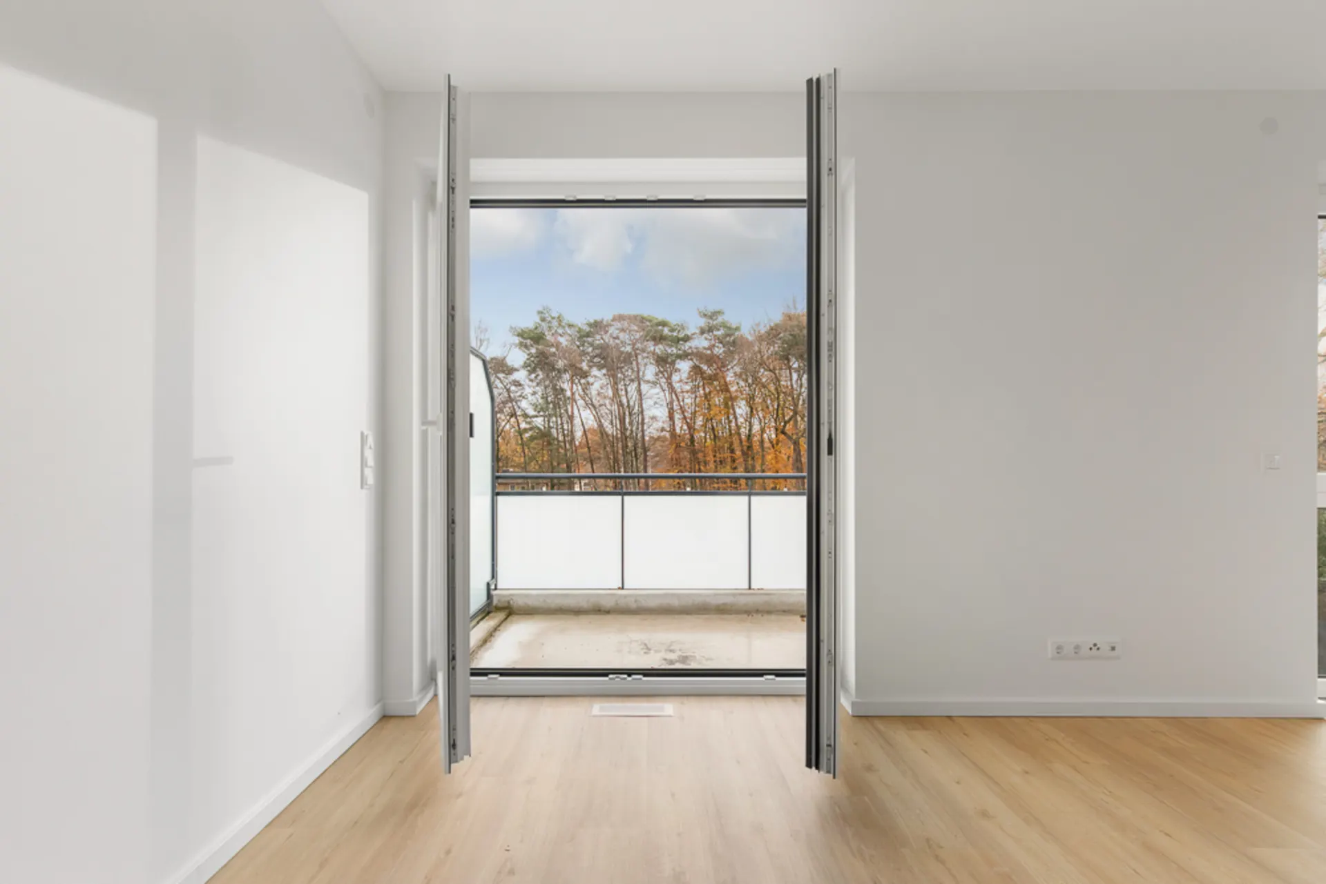 Helles Zimmer mit Holzfußboden und offener Tür zu einem Balkon mit Ausblick auf Bäume.