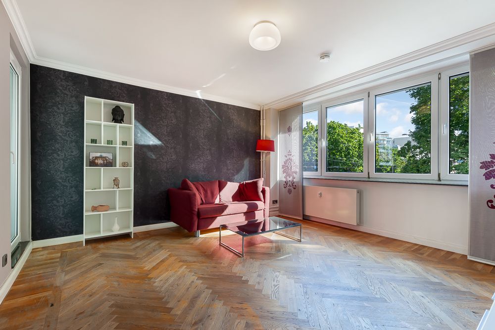 Helles Wohnzimmer mit Parkettboden, rotem Sofa, Glastisch und großem Fenster.