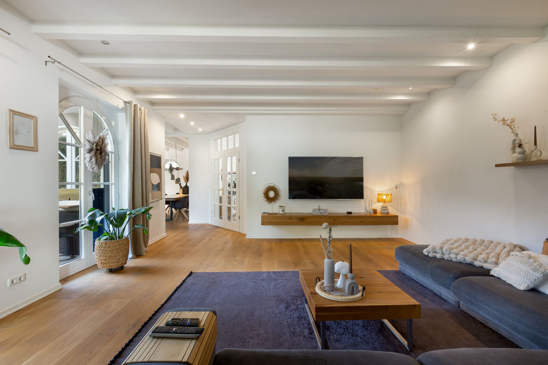 Modernes Wohnzimmer mit grauem Sofa, Holzfußboden, TV an der Wand und Pflanzen am Fenster.