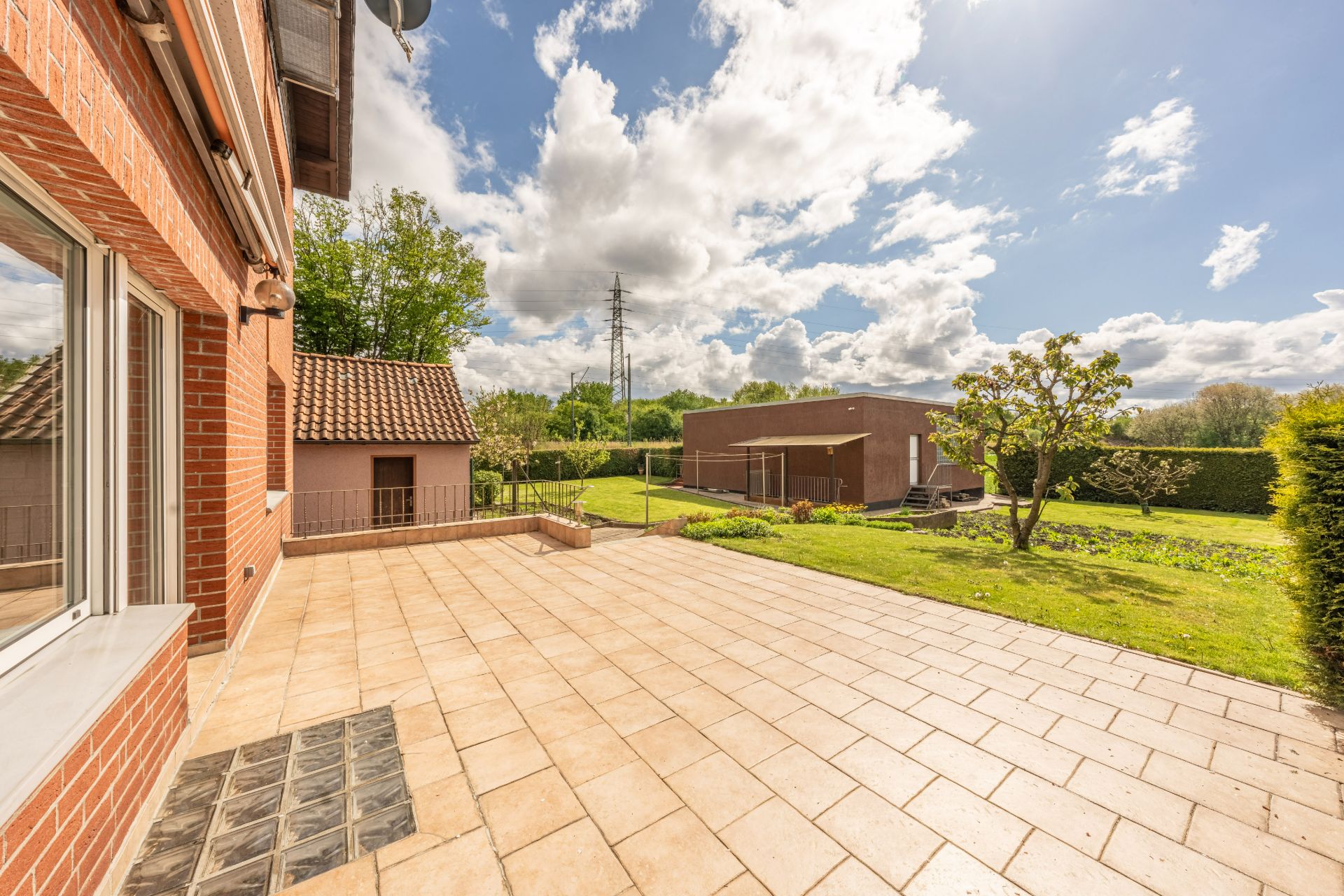 Große geflieste Terrasse mit Blick auf Garten, Baum und angrenzende Gebäude bei Sonnenschein.