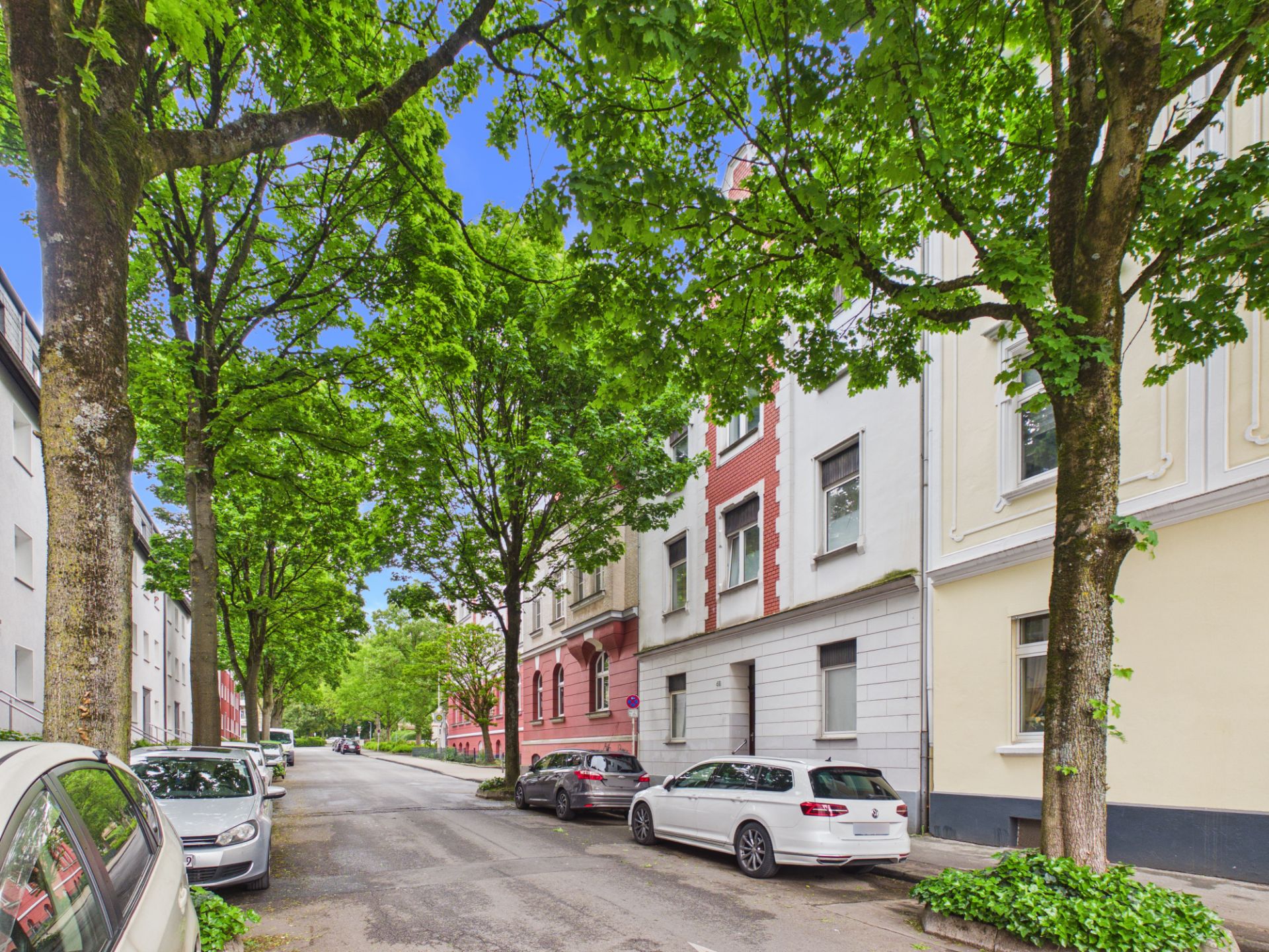Baumbestandene Wohnstraße mit parkenden Autos und mehrstöckigen Wohnhäusern.
