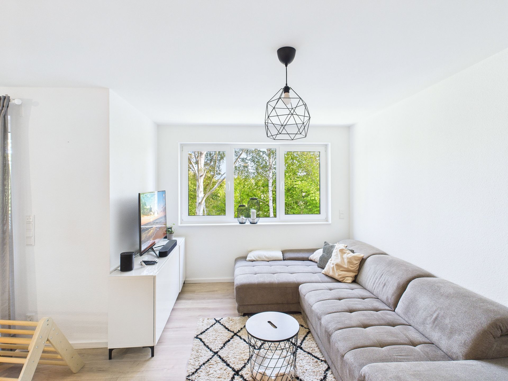 Helles Wohnzimmer mit Ecksofa, Teppich, Couchtisch, TV und großen Fenstern mit Blick ins Grüne.