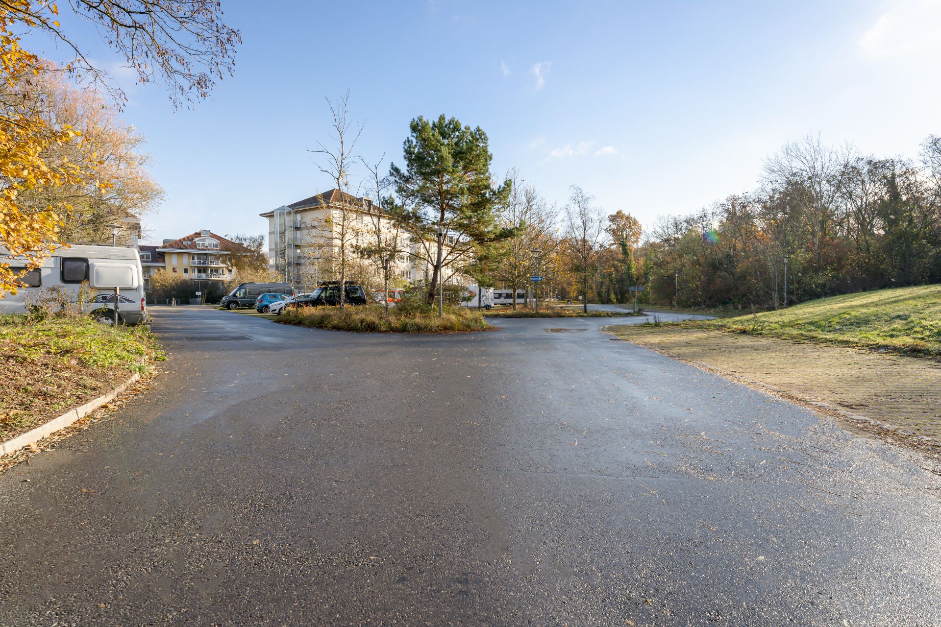 Asphaltierter Parkplatz mit geparkten Autos und Bäumen, Wohngebäude im Hintergrund.