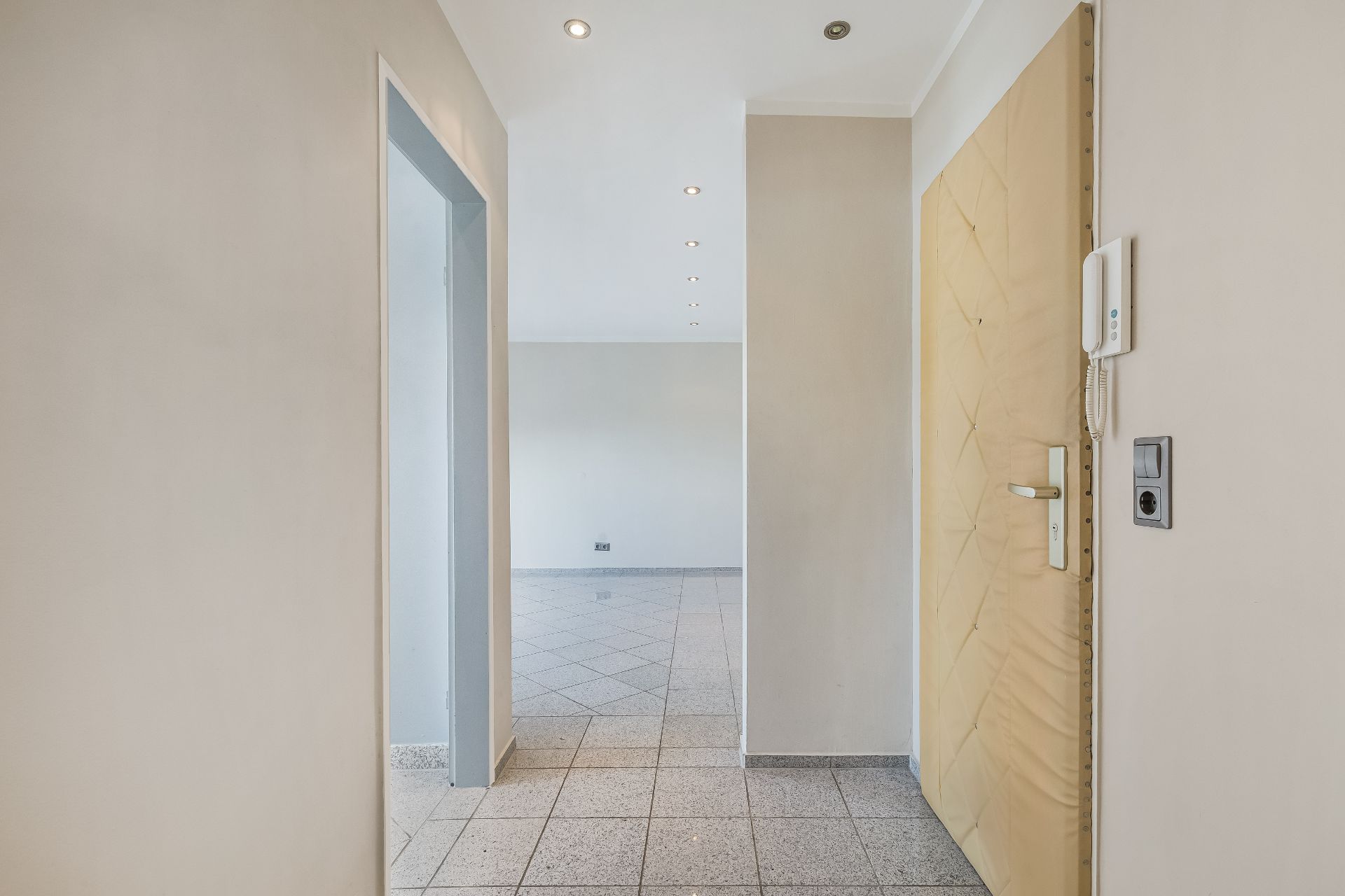 Heller Flur mit Fliesenboden, Tür, Gegensprechanlage und Blick in einen weiteren Raum.