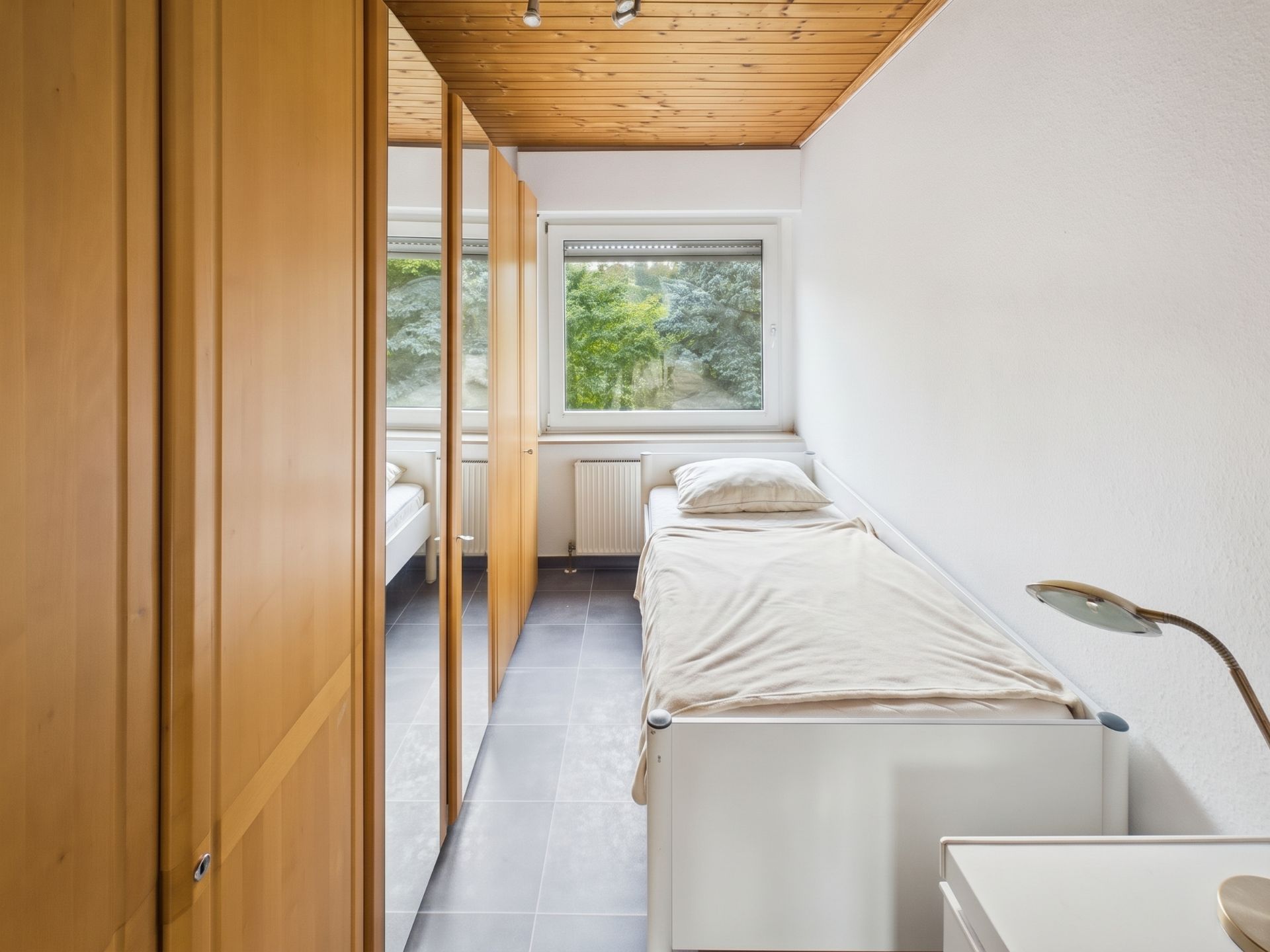 Schmales Schlafzimmer mit Einzelbett, Holzschrank und großem Fenster mit Blick ins Grüne.