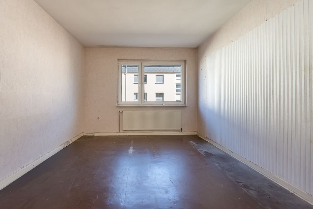 Leerer Raum mit braunem Boden, weiß gestrichenen Wänden und Fenster mit Heizkörper darunter.