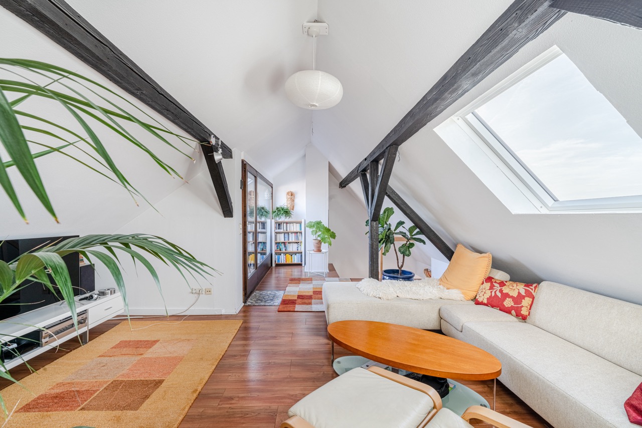 Heller Dachwohnraum mit Sofa, Holztisch, Pflanzen, Teppich und Dachfenster.