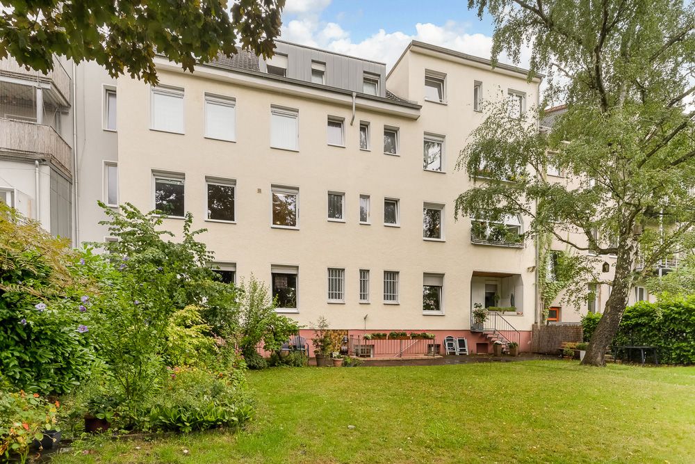 Mehrstöckiges Wohnhaus mit vielen Fenstern, Garten und Bäumen im Vordergrund.