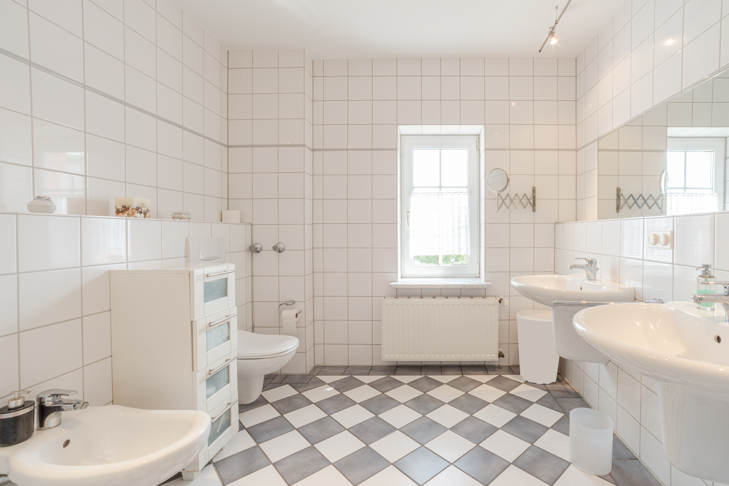 Helles Badezimmer mit zwei Waschbecken, Toilette, Bidet und Fenster, gefliest in Weiß und Grau.