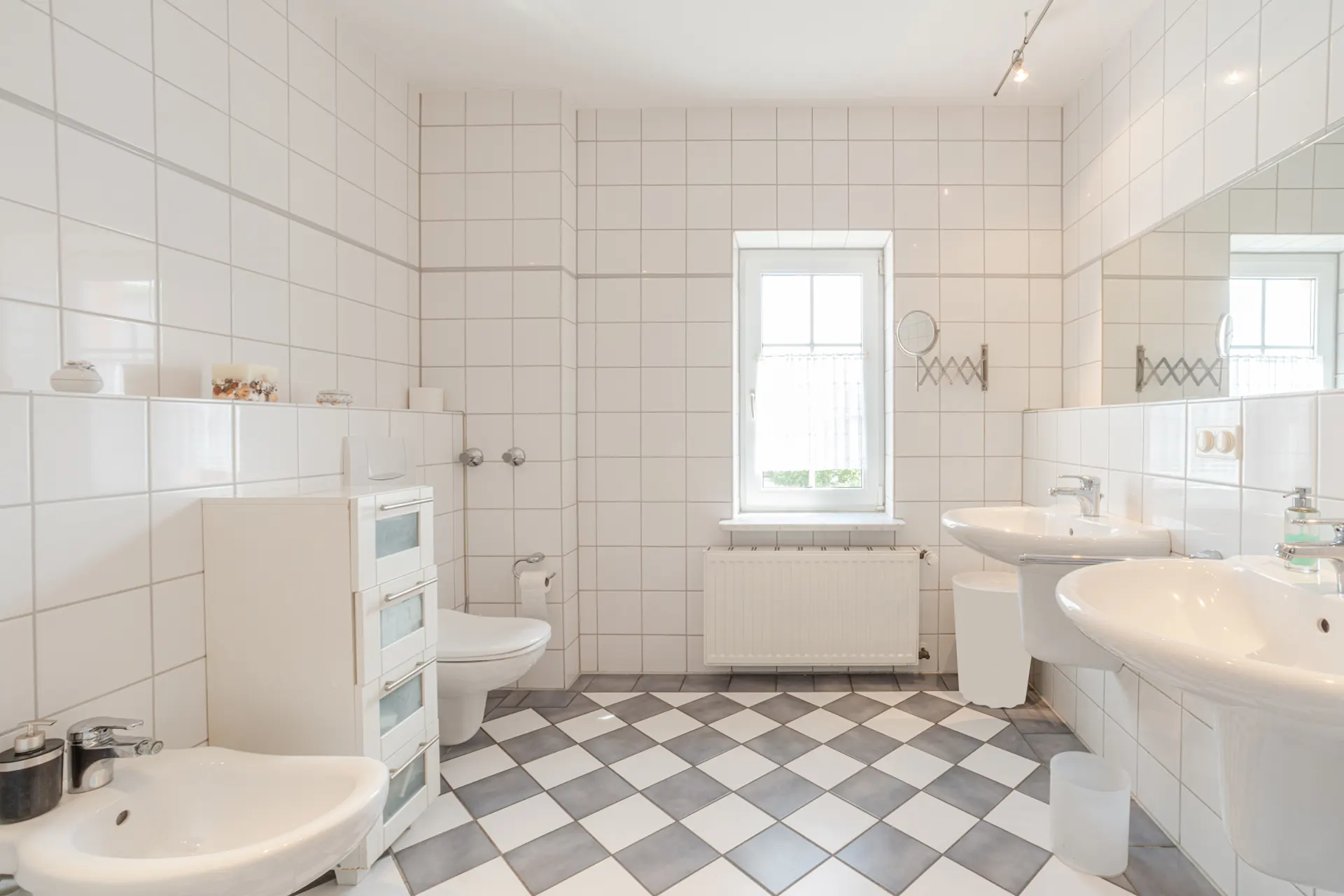 Helles Badezimmer mit zwei Waschbecken, Toilette, Bidet und Fenster, gefliest in Weiß und Grau.