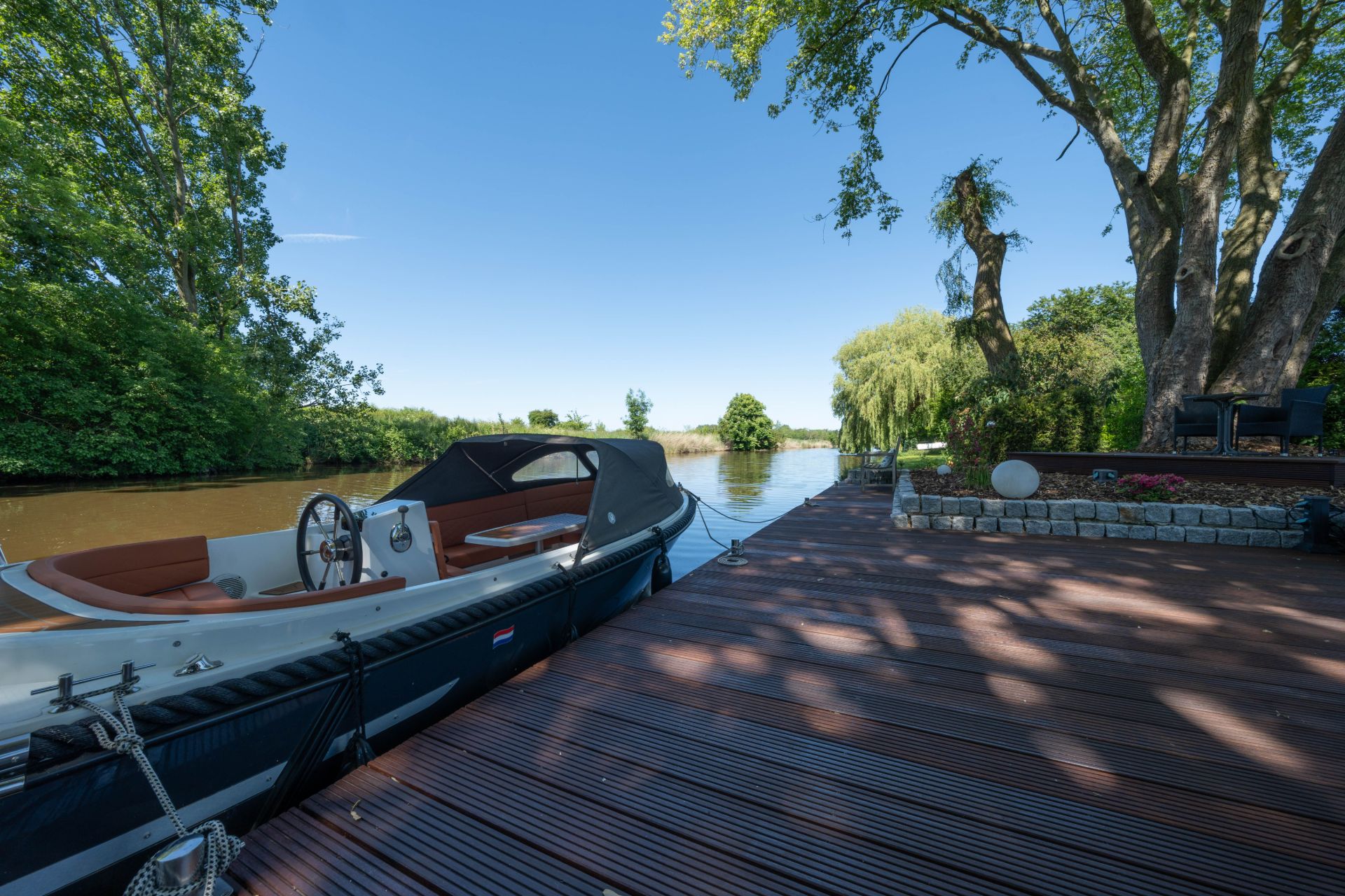 Boot liegt an einem Holzsteg am Fluss, umgeben von Bäumen und Sitzbereich im Garten.