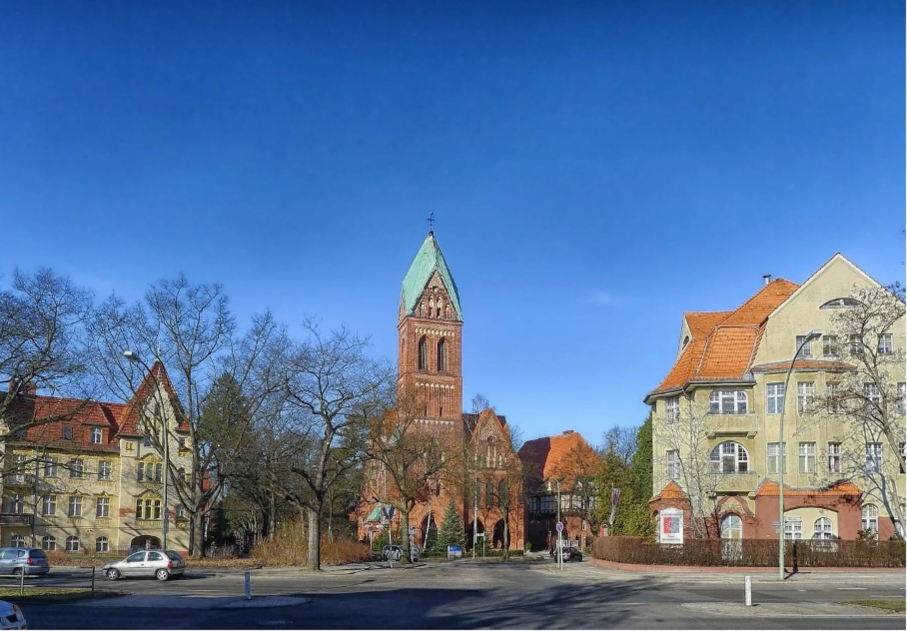 Straßenszene mit Wohnhäusern, Bäumen ohne Laub und einer großen Kirche im Hintergrund.