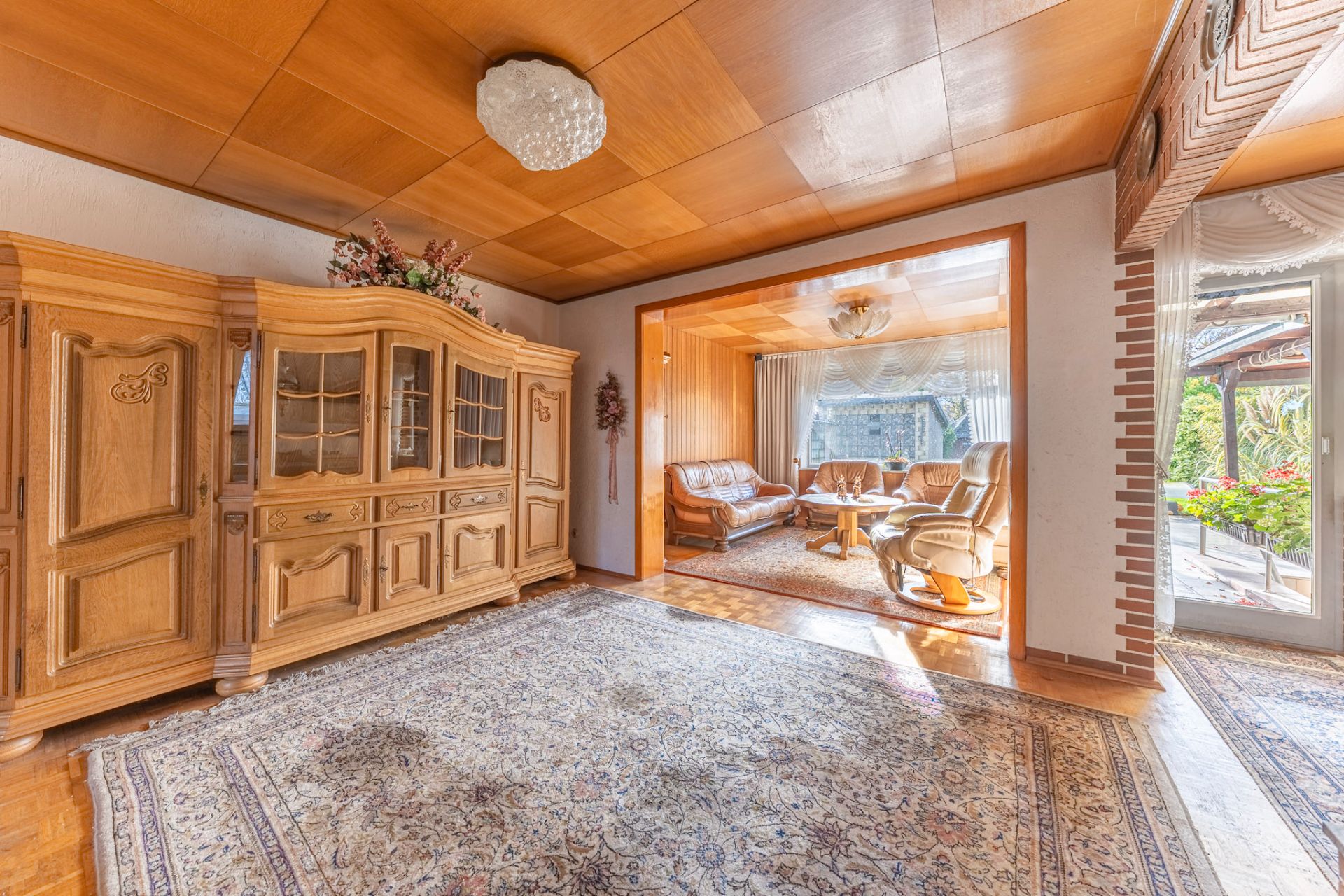 Helles Wohnzimmer mit Teppich, Holzdecke, Schrank, Sitzgruppe und Zugang zur Terrasse.
