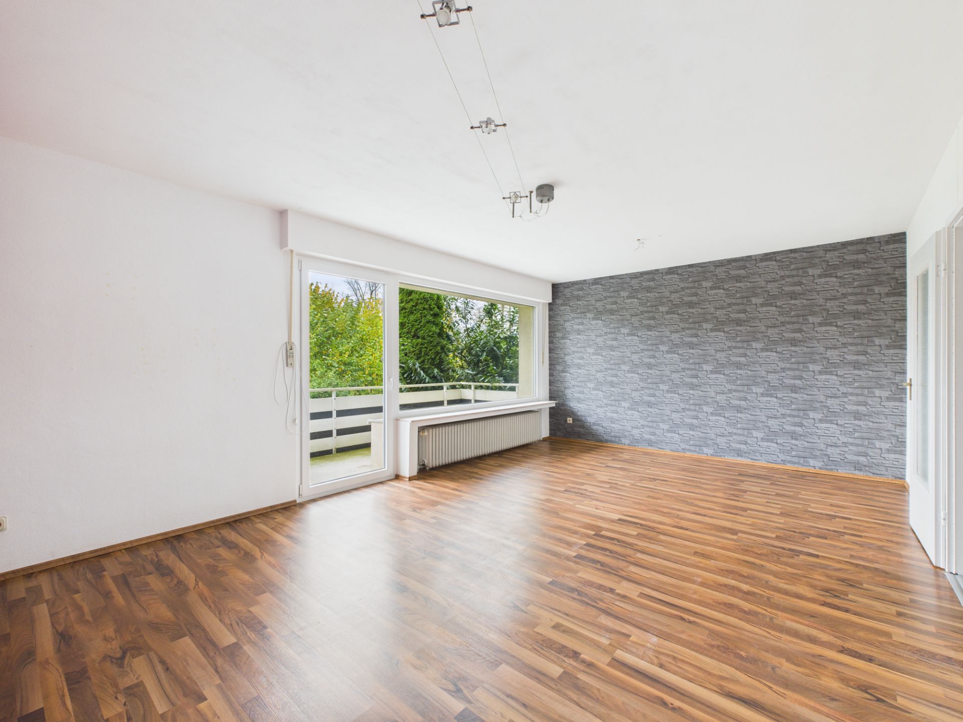 Helles, leeres Zimmer mit Holzfußboden, grauer Steinwand und großer Fensterfront mit Blick ins Grüne.