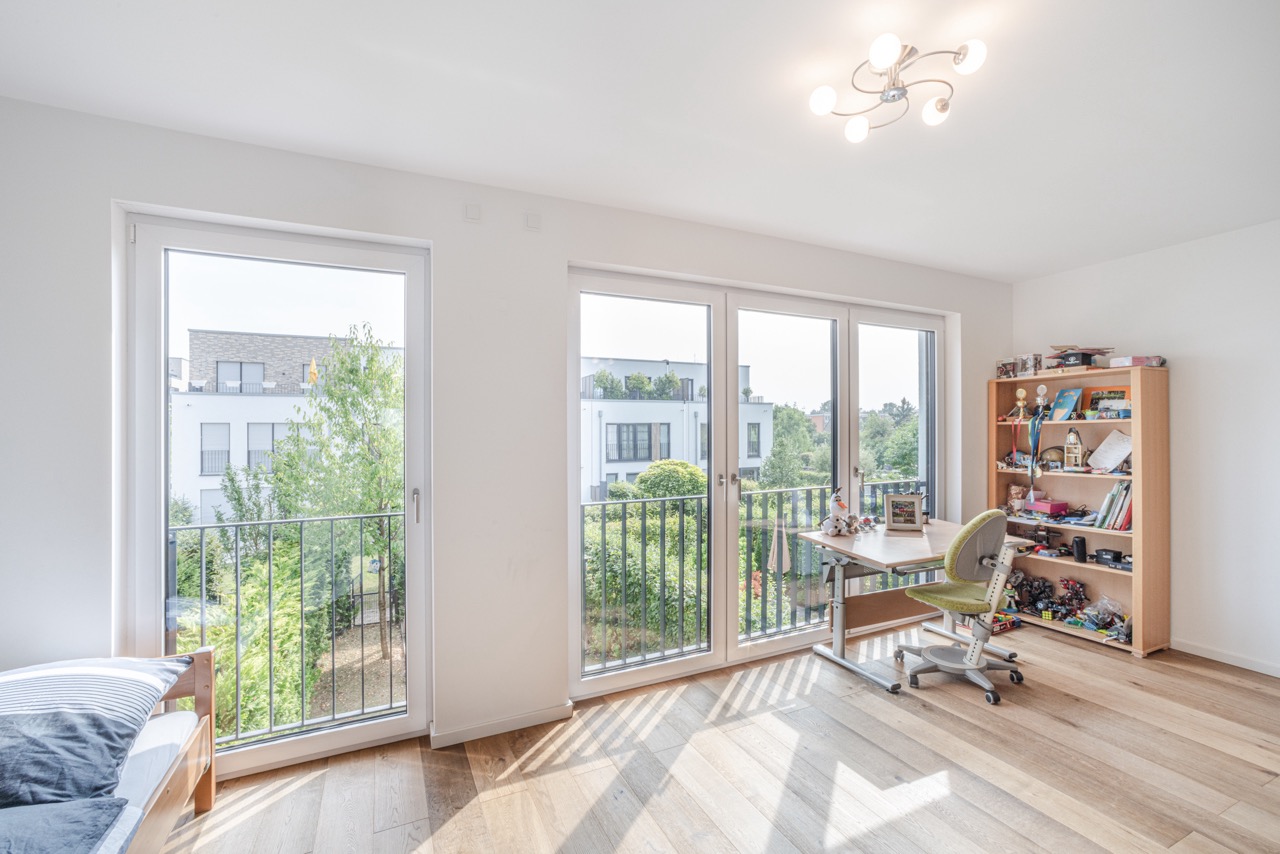 Helles Zimmer mit großen Fenstern, Schreibtisch, Stuhl und Regal mit Spielsachen und Büchern.