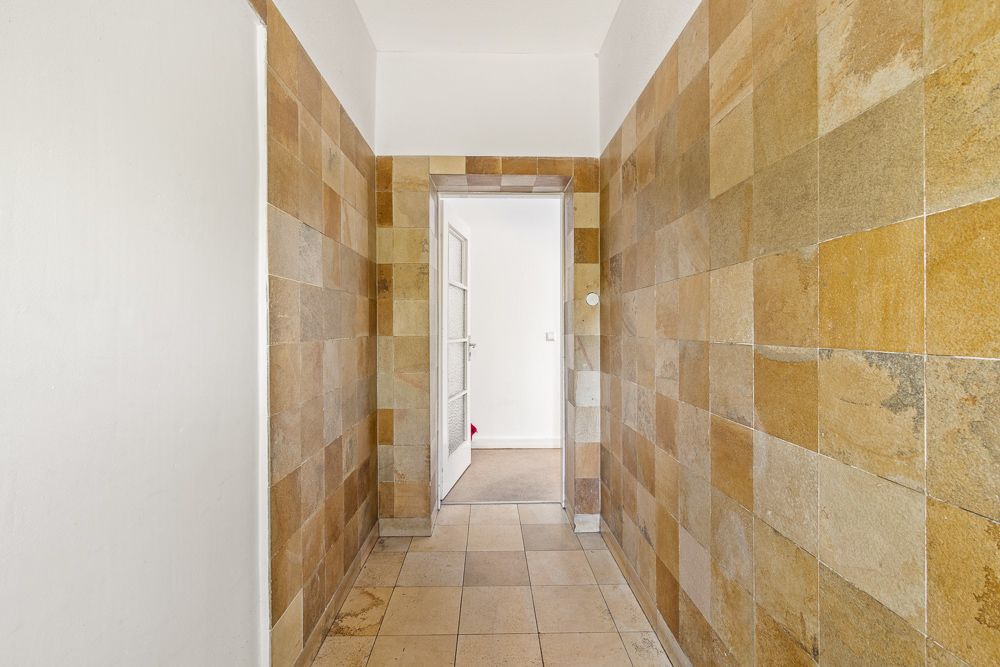 Flur mit beigen Wand- und Bodenfliesen, offene Tür am Ende mit Blick in einen hellen Raum.