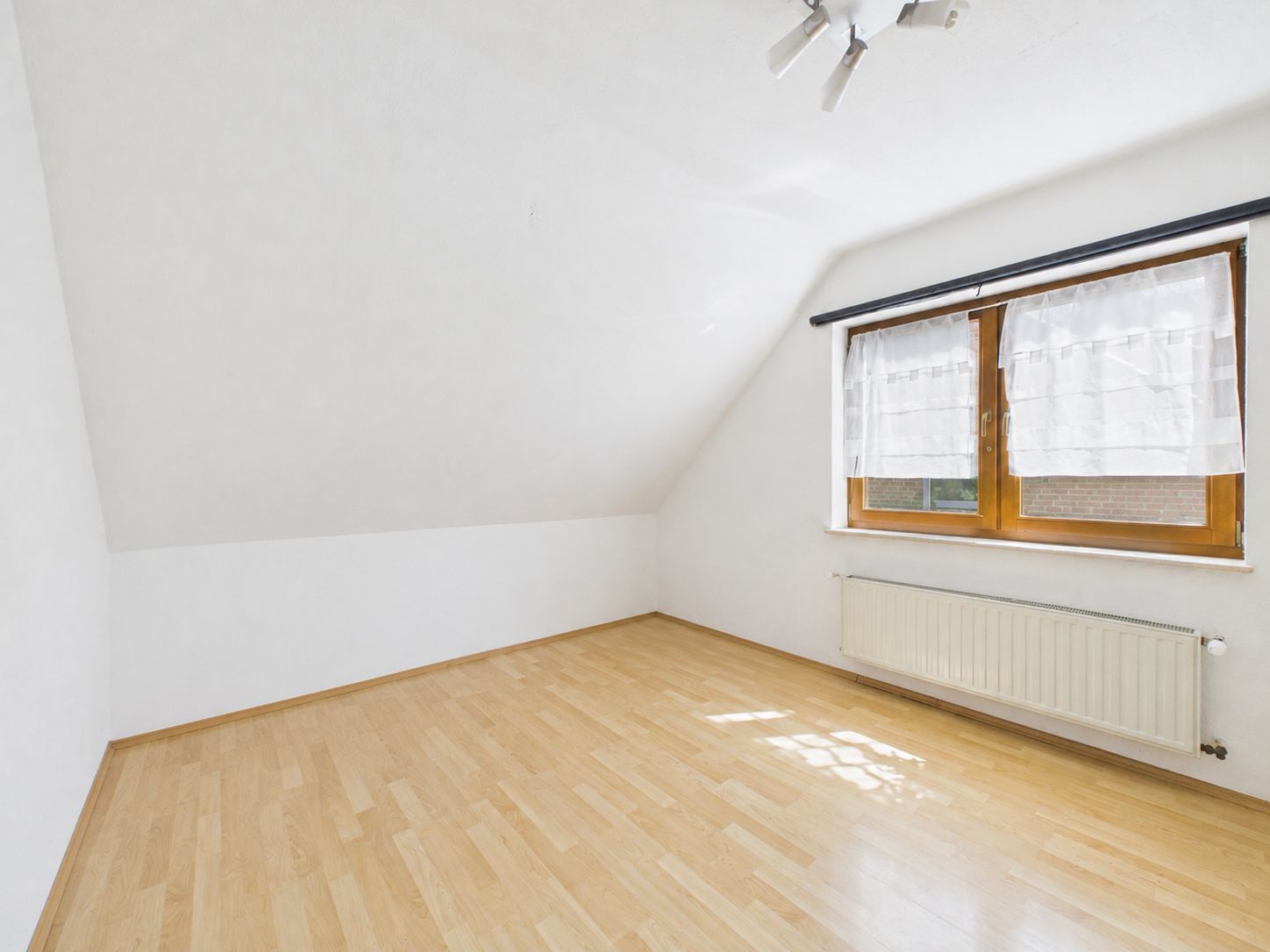 Helles, leeres Zimmer mit schräger Decke, großem Fenster und Parkettboden.