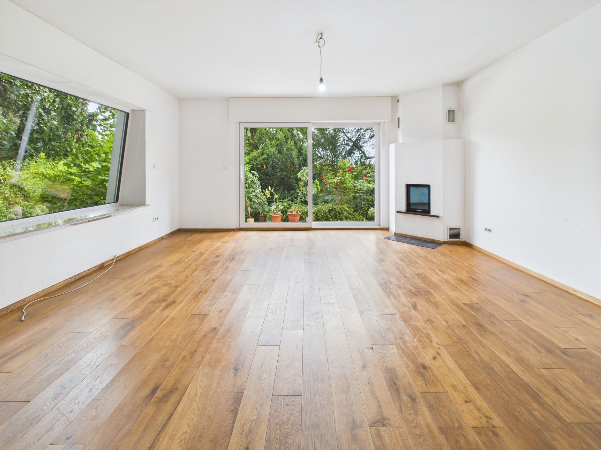 Leeres Wohnzimmer mit Holzboden, großem Fenster, Kamin und Zugang zur Terrasse.
