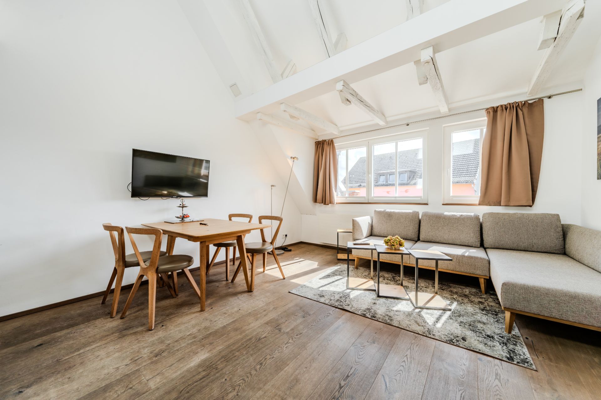Helles Wohnzimmer mit Ecksofa, Esstisch, Stühlen, Wandfernseher und großen Fenstern.