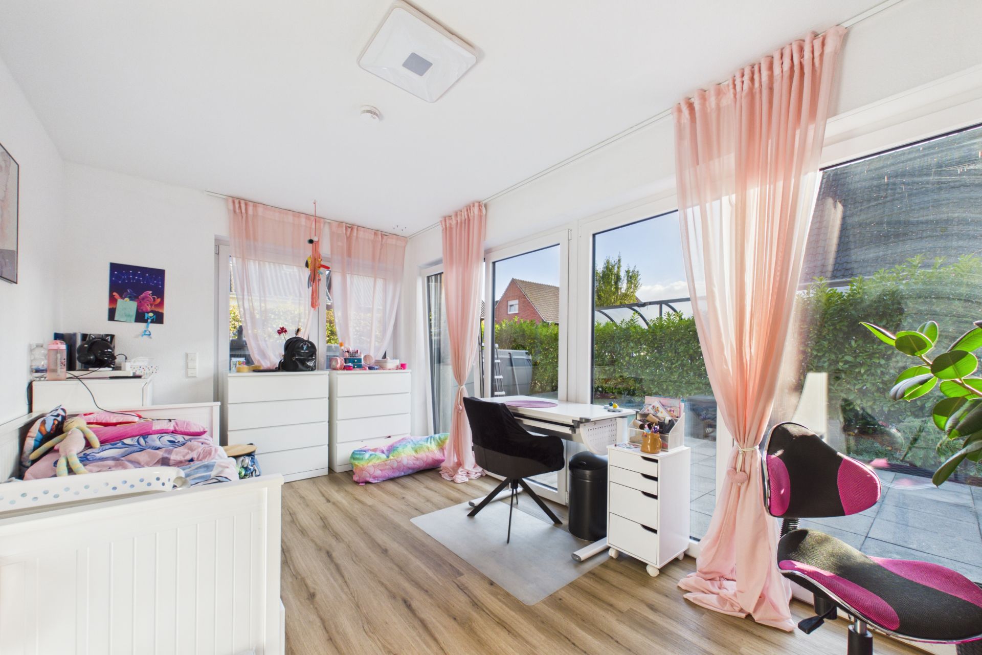 Helles Kinderzimmer mit Schreibtisch, Bett, zwei Kommoden und rosa Vorhängen vor großen Fenstern.