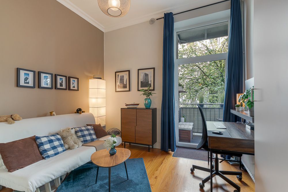 Wohnzimmer mit Sofa, Couchtisch, Schreibtisch und Zugang zu einem Balkon mit Blick auf Bäume.