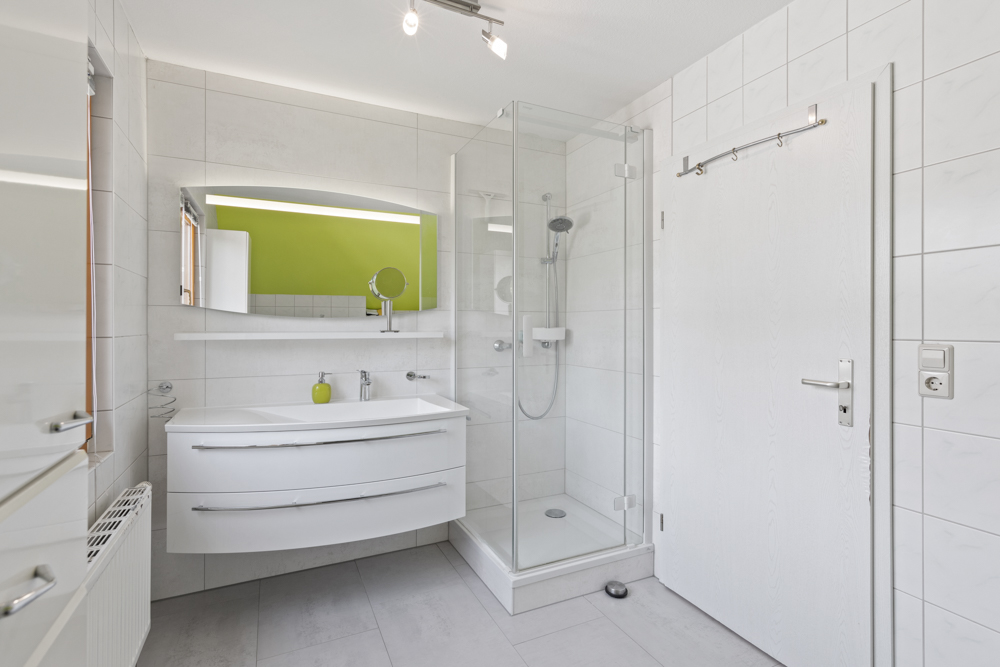 Modernes Badezimmer mit Dusche, weißem Waschbecken und großem Spiegel, grüne Wand im Hintergrund.
