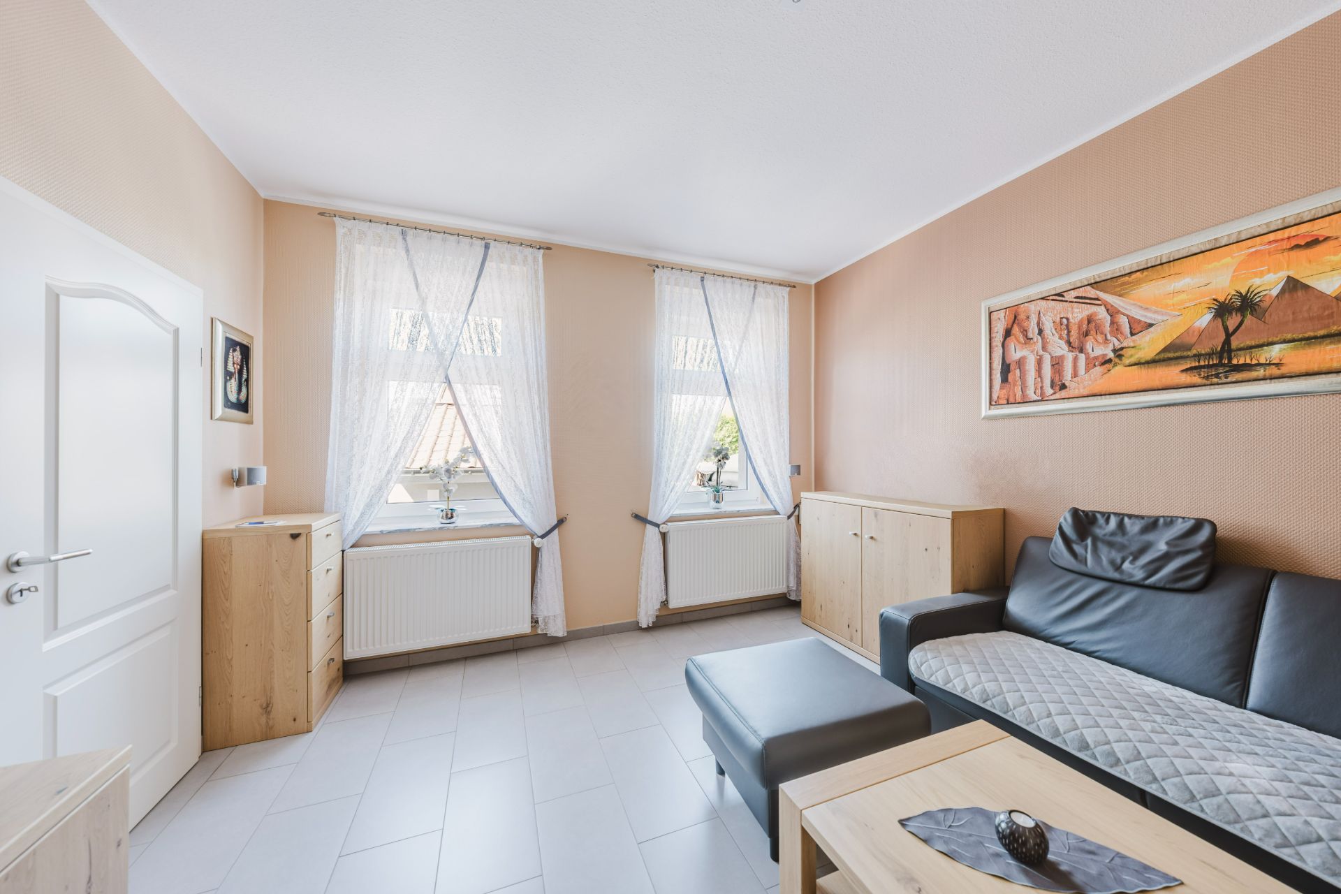 Helles Wohnzimmer mit Sofa, Couchtisch, zwei Fenstern und Wandbild mit Wüstenmotiv.