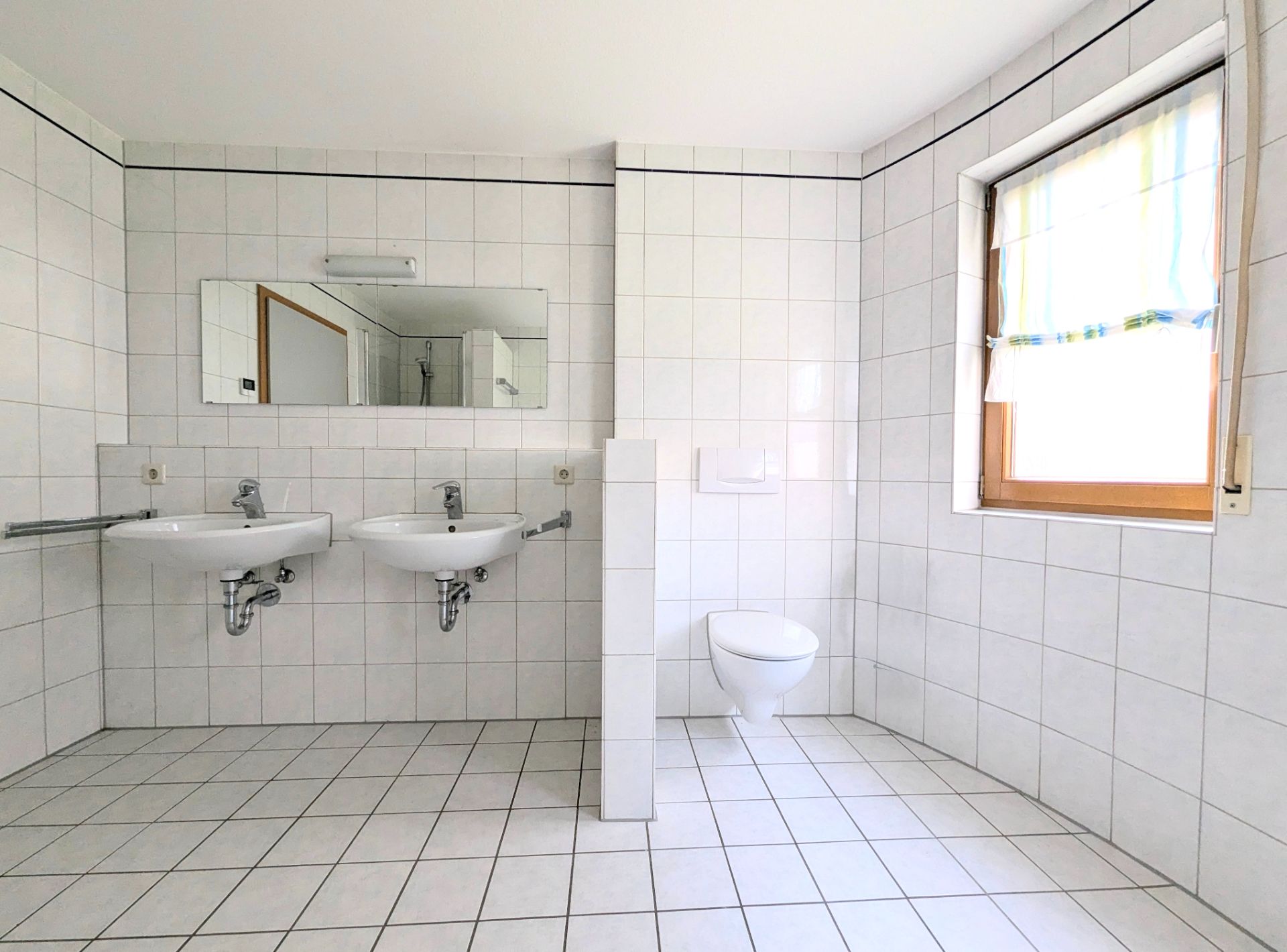 Helles Badezimmer mit zwei Waschbecken, Wandspiegel, WC und Fenster mit Rollovorhang.
