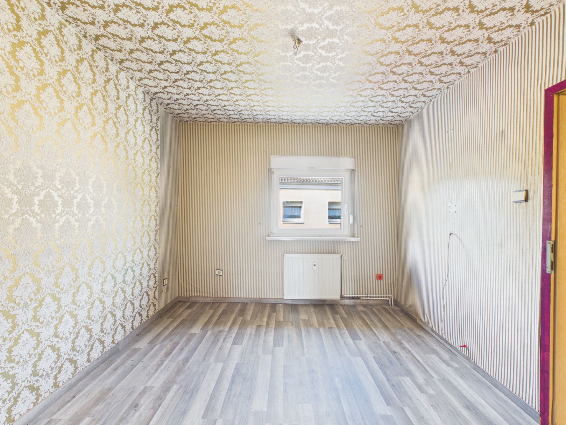 Leerer Raum mit gemusterten Tapeten, Fenster und grauem Laminatboden.