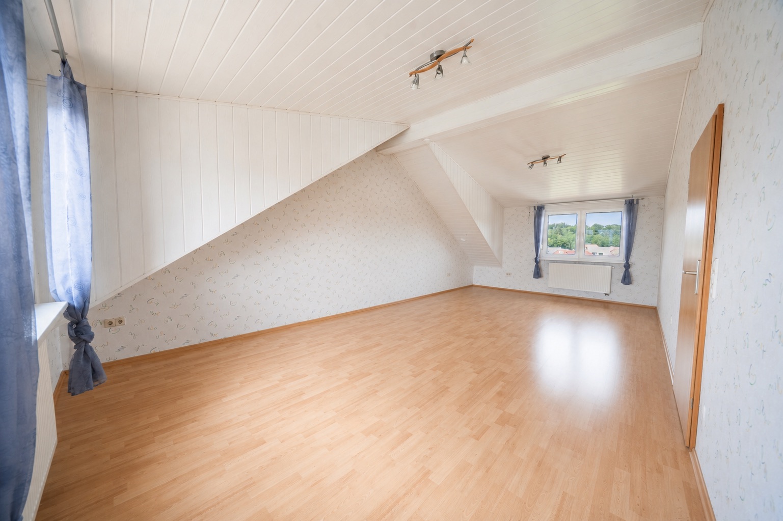 Helles, leeres Dachgeschosszimmer mit Holzfußboden, Dachschräge und zwei Fenstern mit blauen Vorhängen.