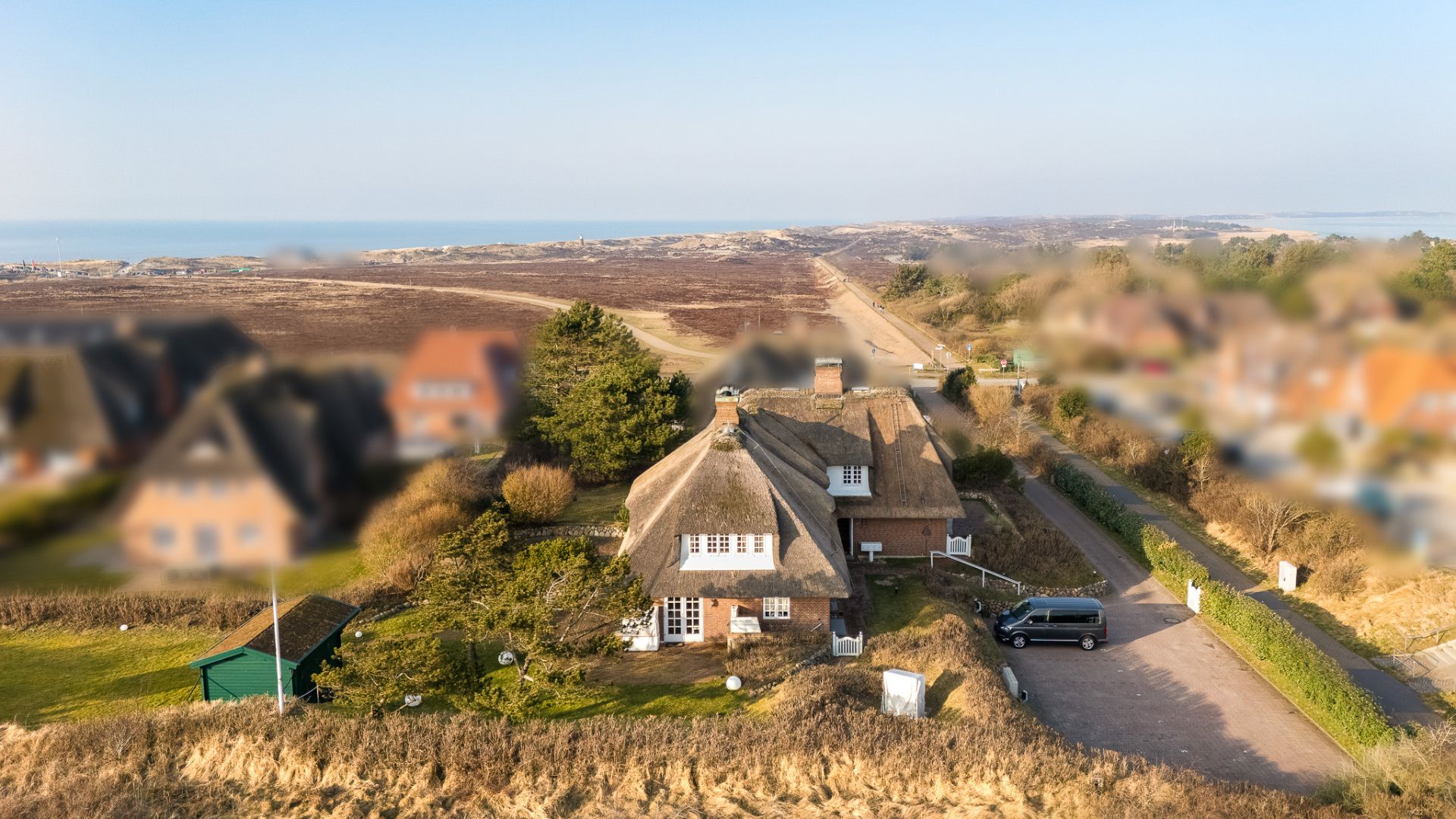 Reetgedecktes Haus mit Garten, daneben eine Einfahrt mit Auto, umgeben von Dünenlandschaft.