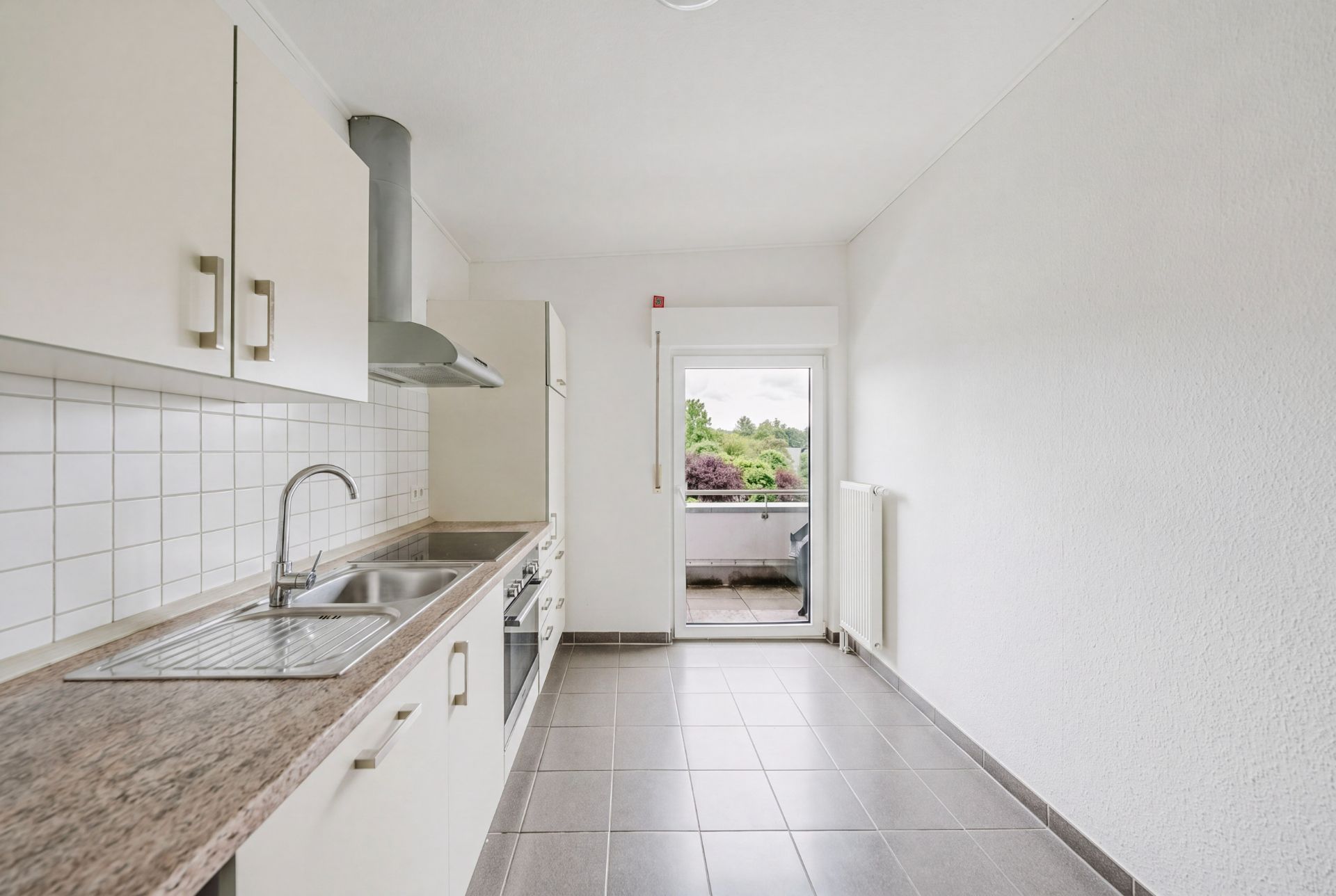 Helle Einbauküche mit Fliesenboden und Zugang zu einem Balkon mit Ausblick ins Grüne.