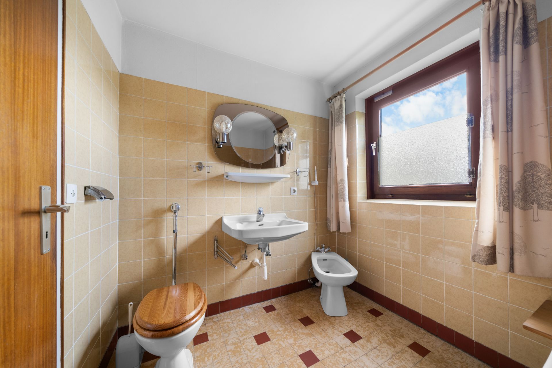 Badezimmer mit WC, Bidet, Waschbecken, Spiegel und Fenster mit Vorhang.