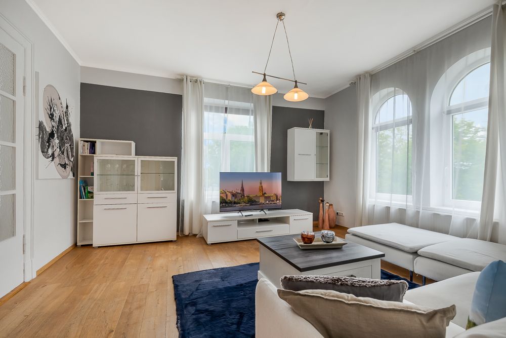 Helles Wohnzimmer mit Sofa, Couchtisch, Fernseher, Sideboard und großen Fenstern mit Vorhängen.