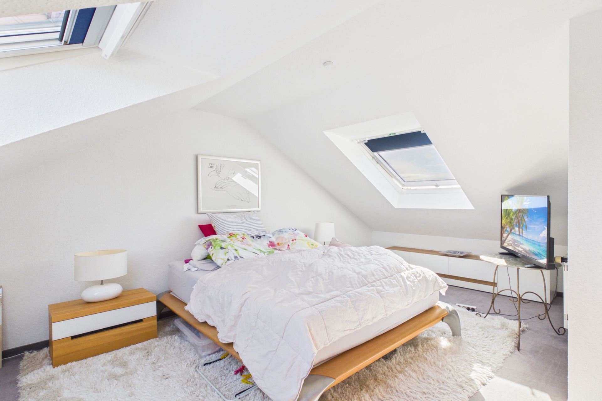 Helles Schlafzimmer mit Dachfenster, Doppelbett, Nachttisch und Fernseher an der Wand.
