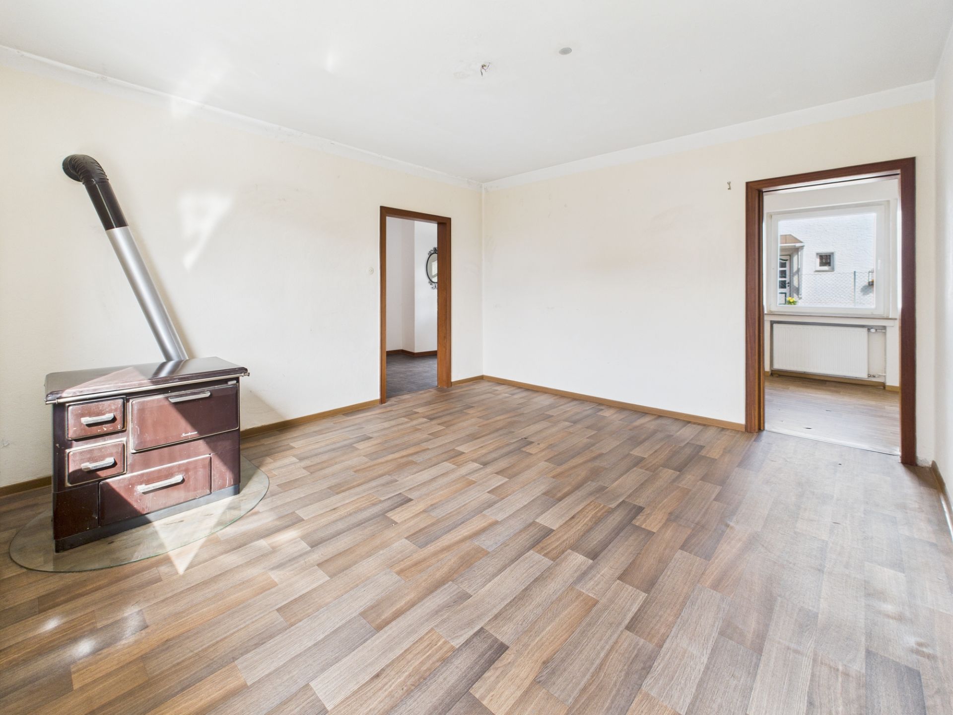 Helles Wohnzimmer mit Holzofen, Laminatboden und zwei offenen Türrahmen.