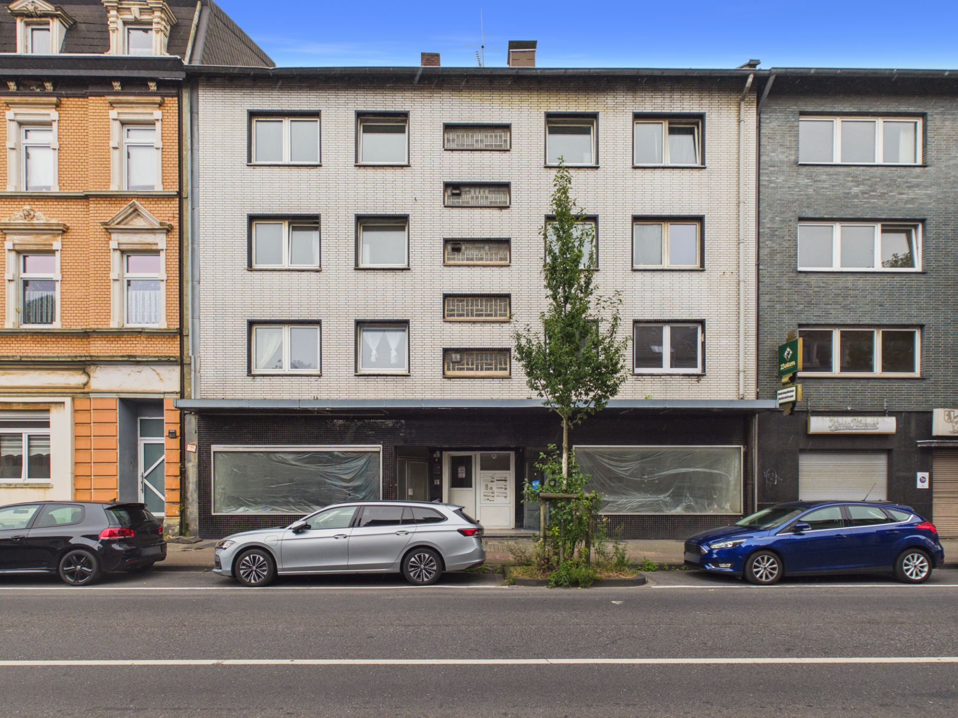 Vierstöckiges Wohnhaus mit weißer Fassade, zwei Schaufenstern im Erdgeschoss und Autos vor dem Gebäude.