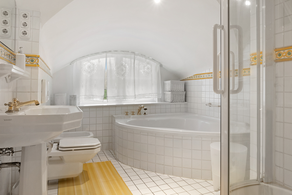 Helles Badezimmer mit Eckbadewanne, Dusche, Waschbecken und WC unter einem Rundfenster.